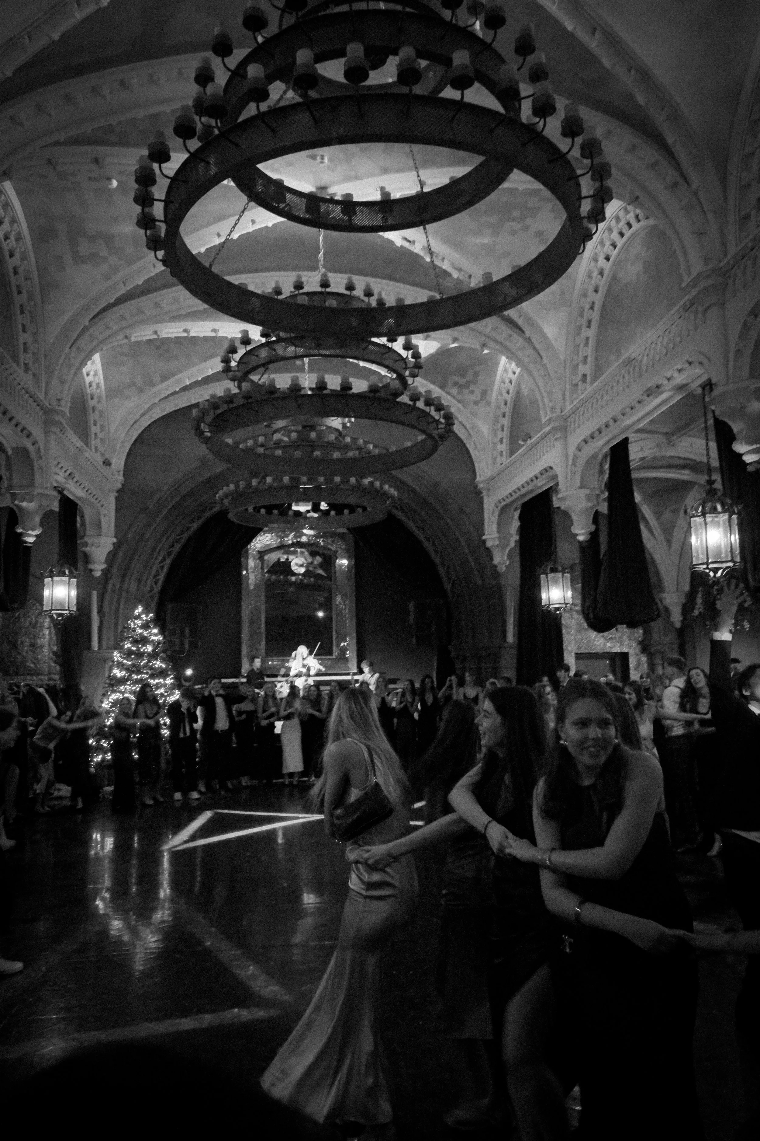 A black-and-white photo of a dance hall with a high, arched ceiling decorated with circular chandeliers. People, mostly young women, are dancing in pairs, holding hands and enjoying the music. A band or musicians are performing on a stage at the back