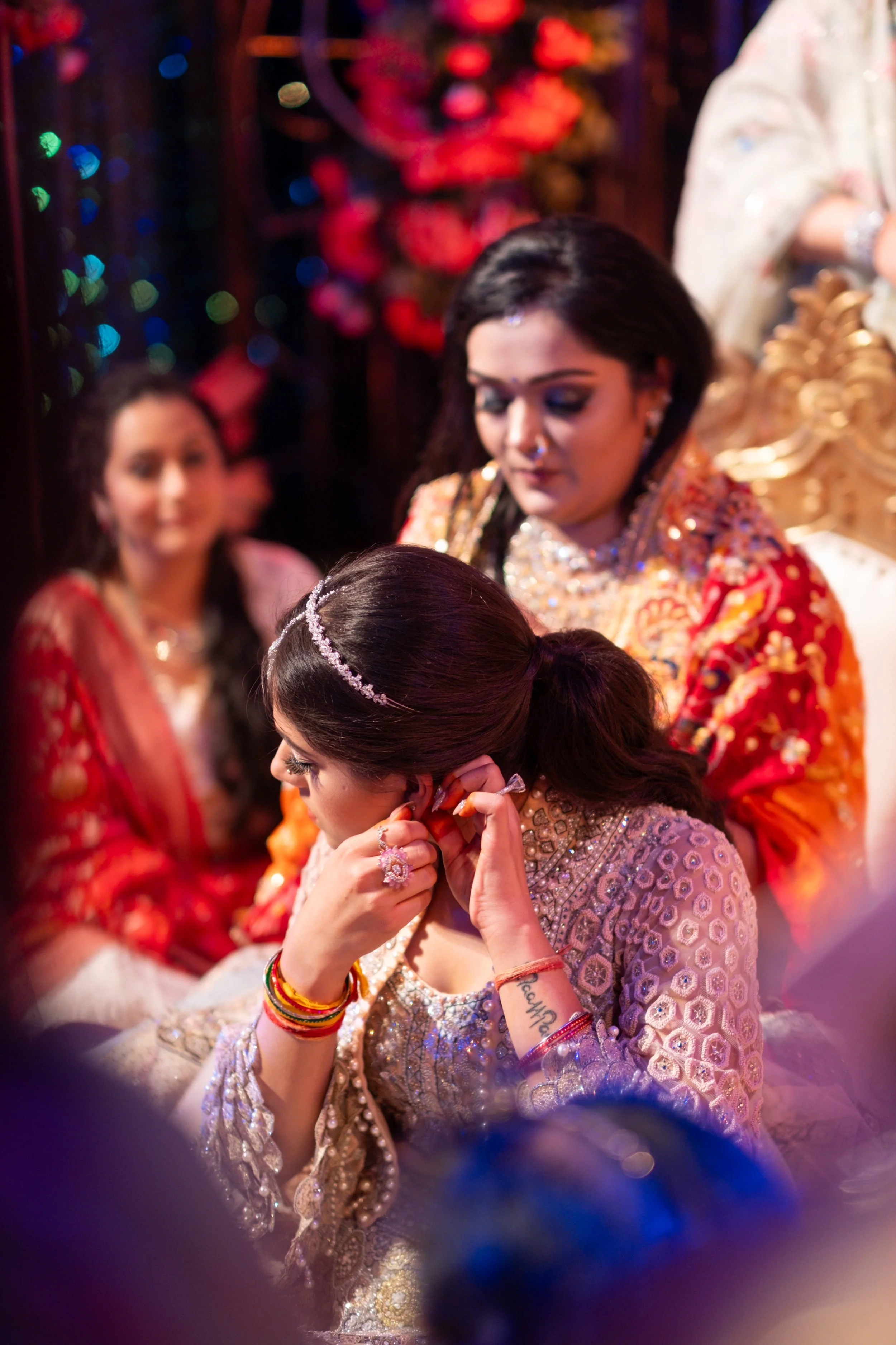 A woman dressed in an ornate, traditional outfit is adjusting her earrings at a cultural or wedding event. She is adorned with jewelry and has a tattoo on her wrist. Behind her, another woman in a red sari and jewelry is visible, and in the backgroun