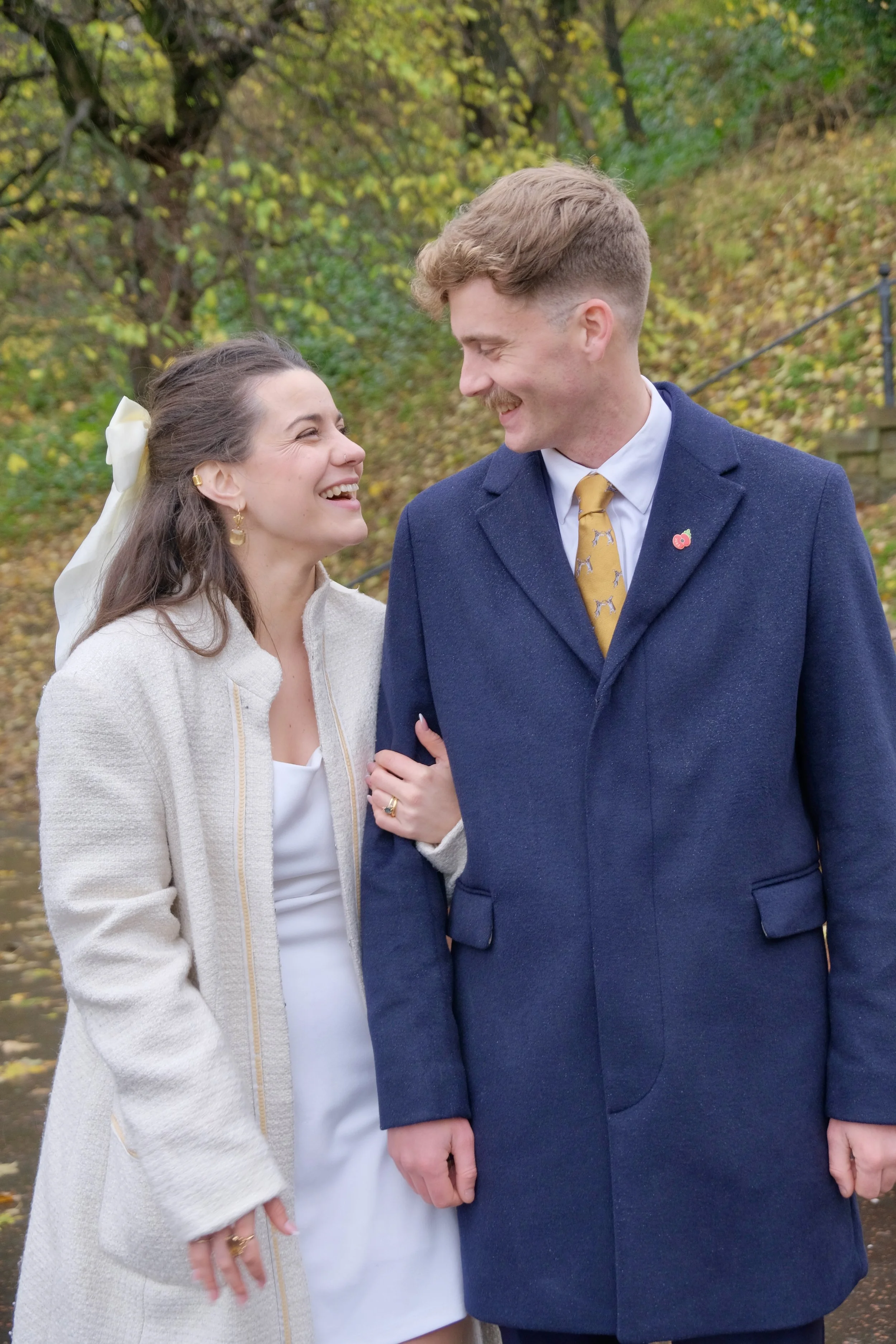 A couple smiling and looking at each other outdoors during fall, with trees and fallen leaves in the background.