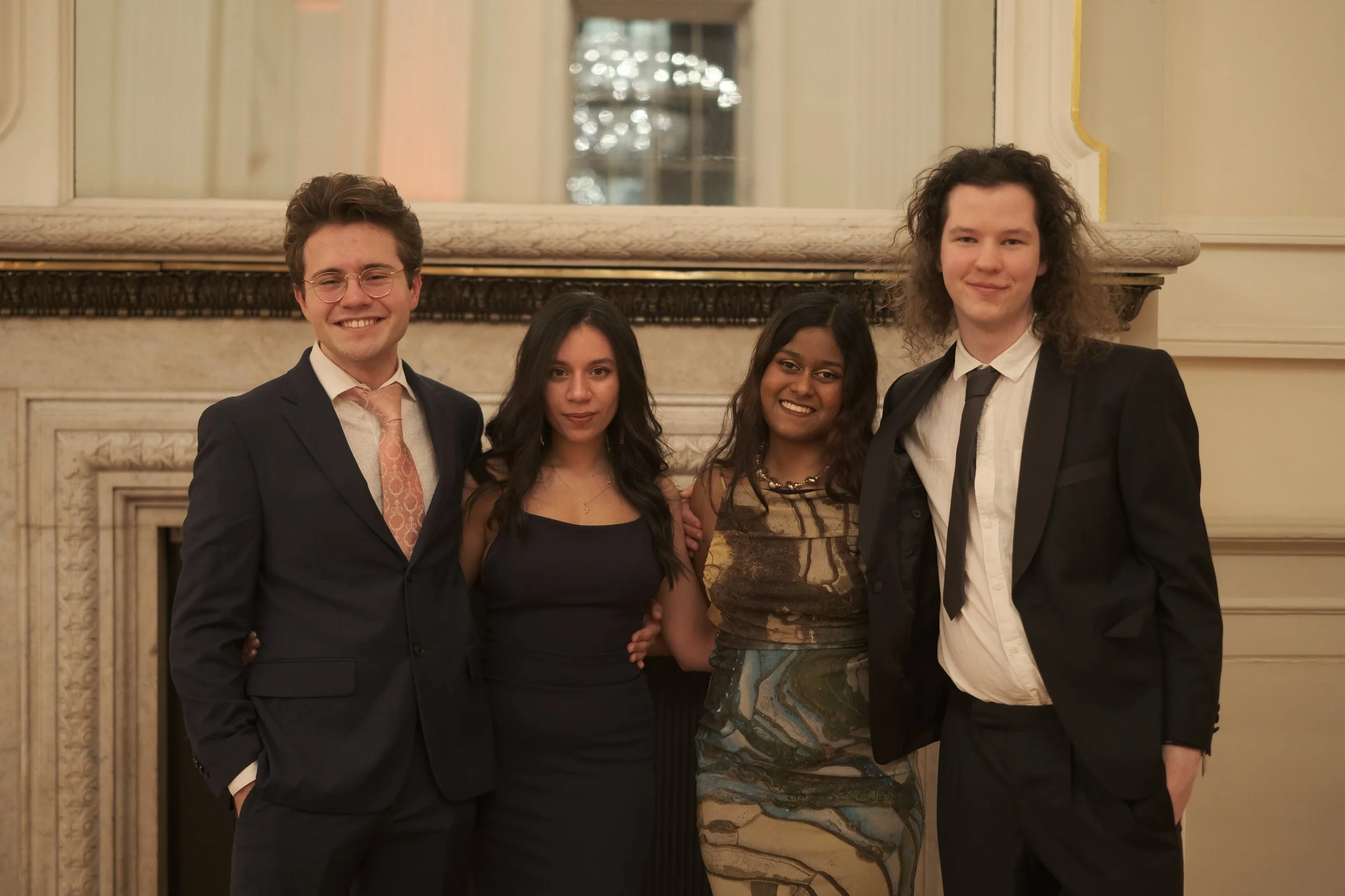 Four young adults dressed in formal attire, standing together in front of a fireplace, smiling at an indoor event.