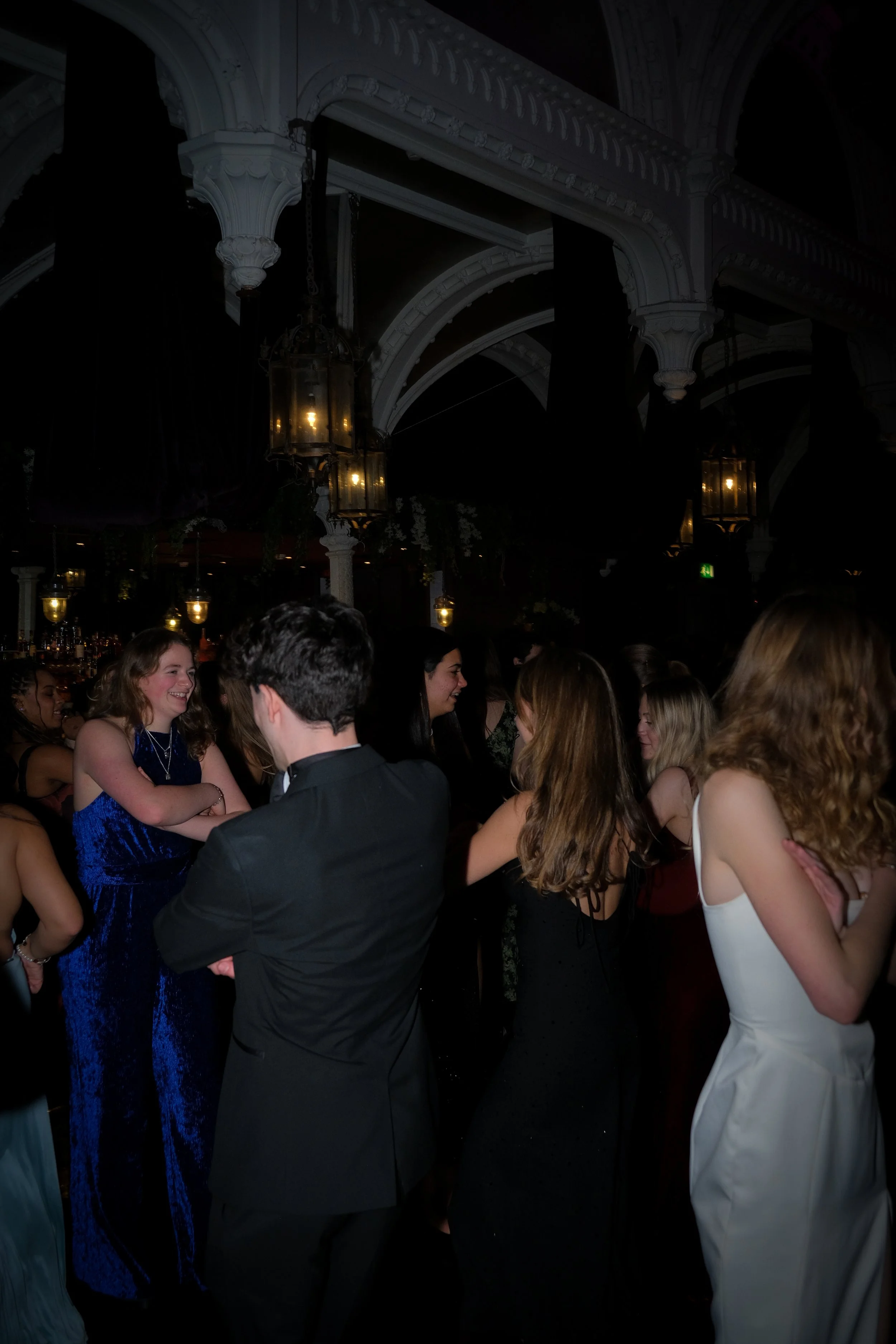 People socializing and laughing at a formal event or party in a dimly lit venue with ornate architectural details and hanging lantern-style lights.