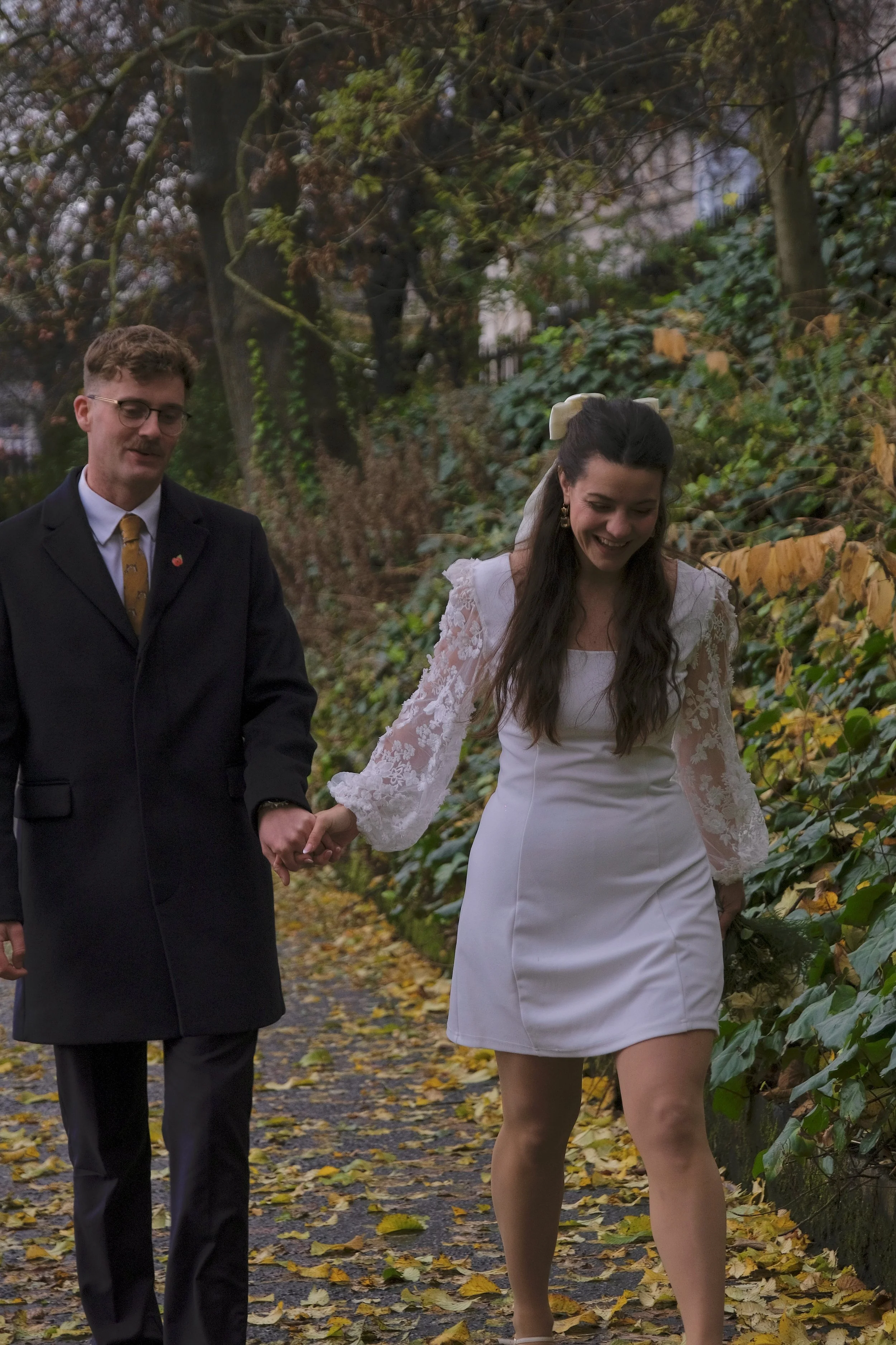 A young woman in a white dress with lace sleeves and a large ribbon in her long dark hair is walking hand-in-hand with a man in a black suit and glasses on a leaf-covered pathway surrounded by trees and bushes during autumn.