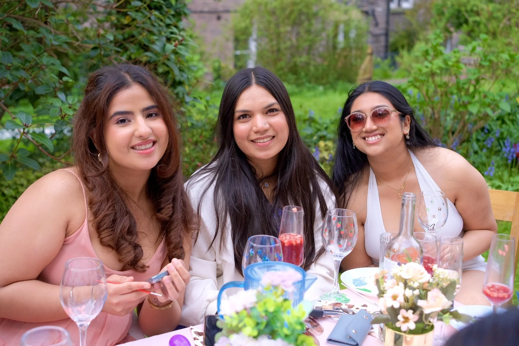 Three women sitting at a dining table outdoors, smiling, surrounded by flowers and glasses of drinks.