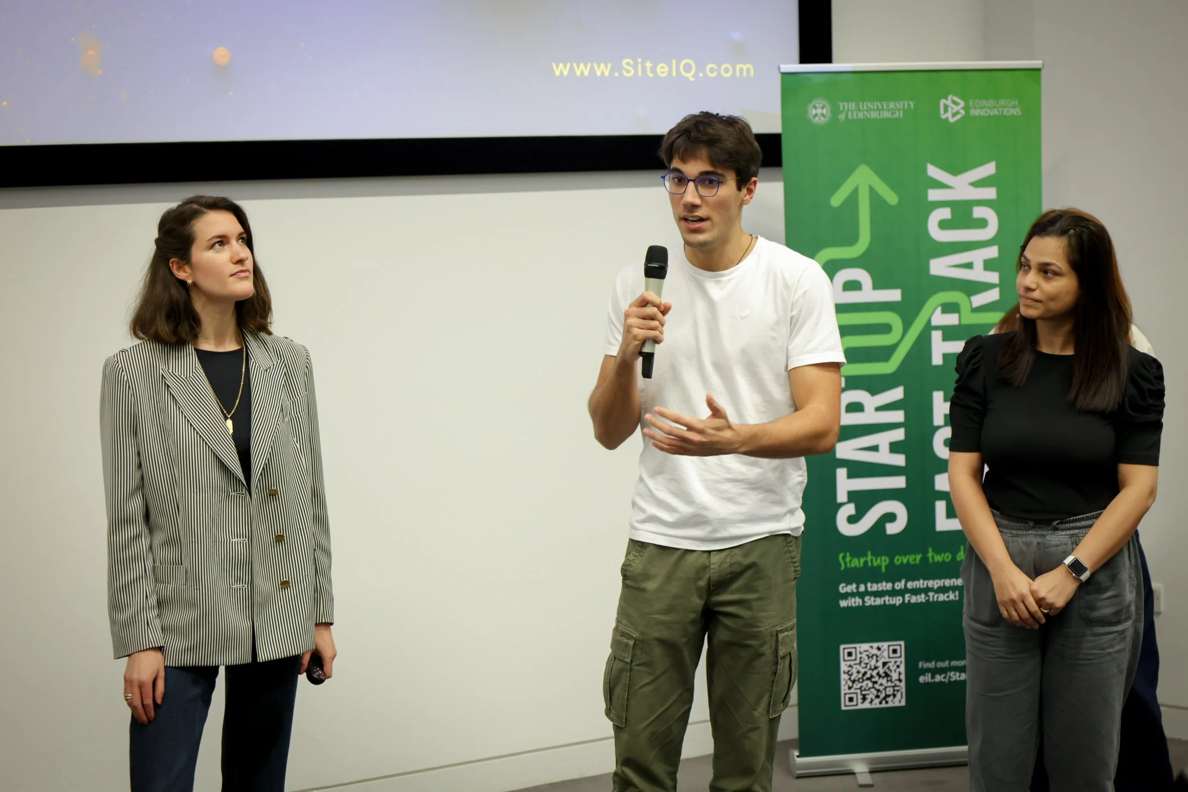A man in a white t-shirt speaking into a microphone during a presentation, flanked by two women, one wearing a striped blazer and the other in a black top, in front of a green banner with upward arrows and startup-related text.