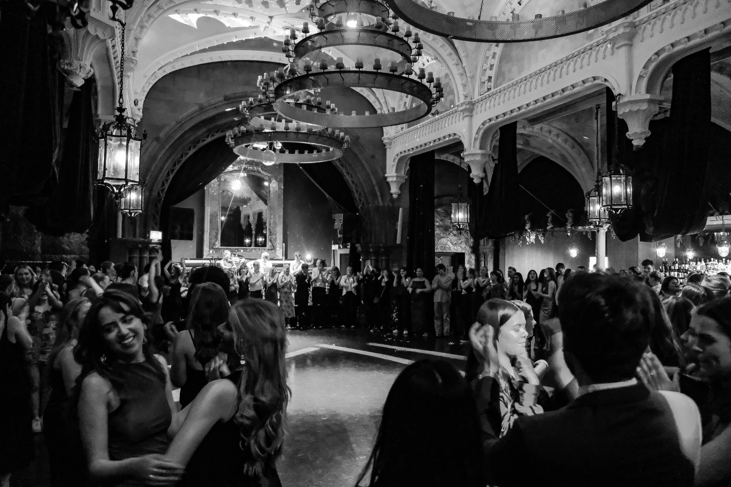 People dancing and socializing in a ballroom with ornate chandeliers and a stage in the background.