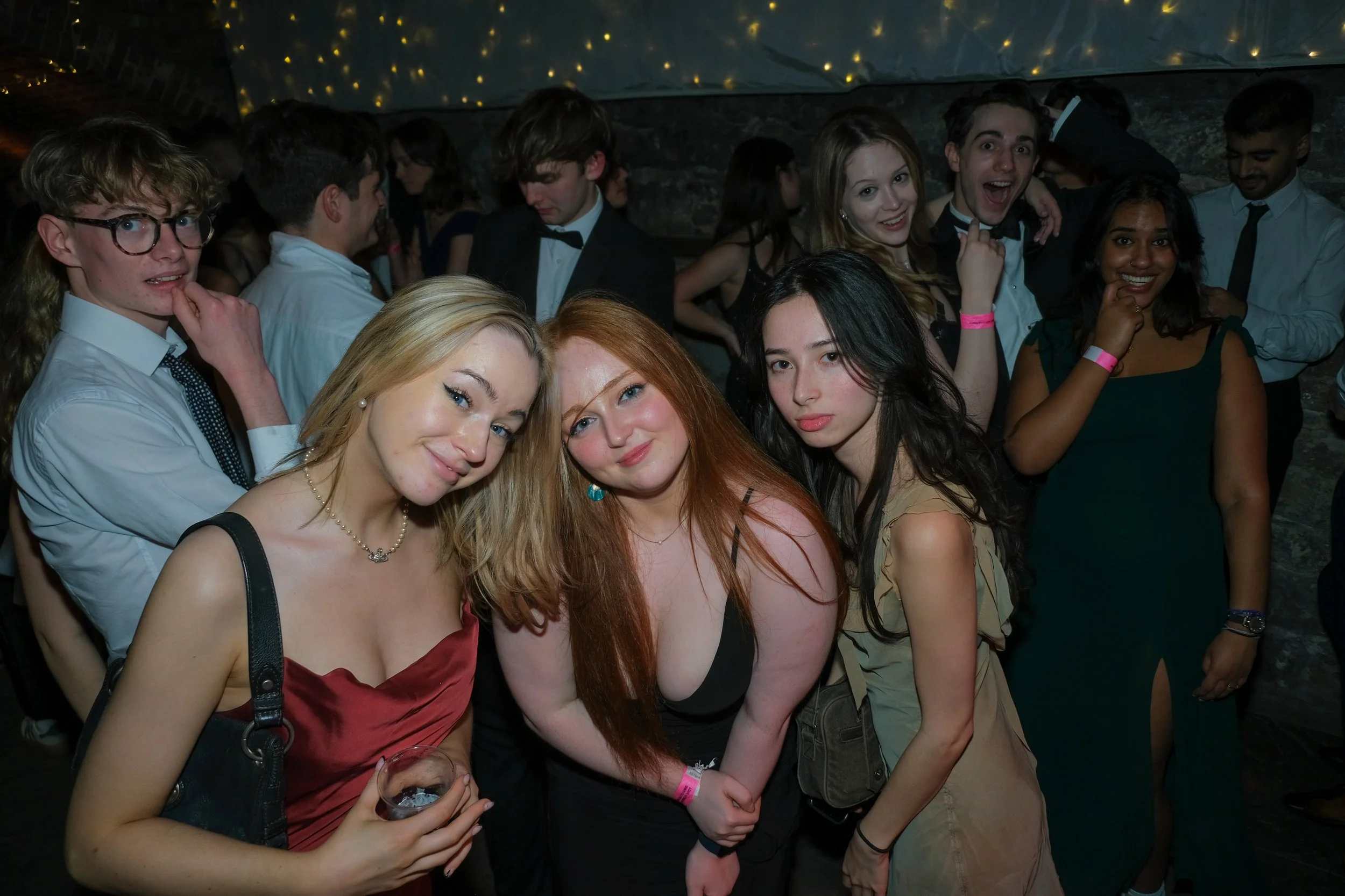 Group of young people at a party or event, some smiling and others looking at the camera, with fairy lights overhead.
