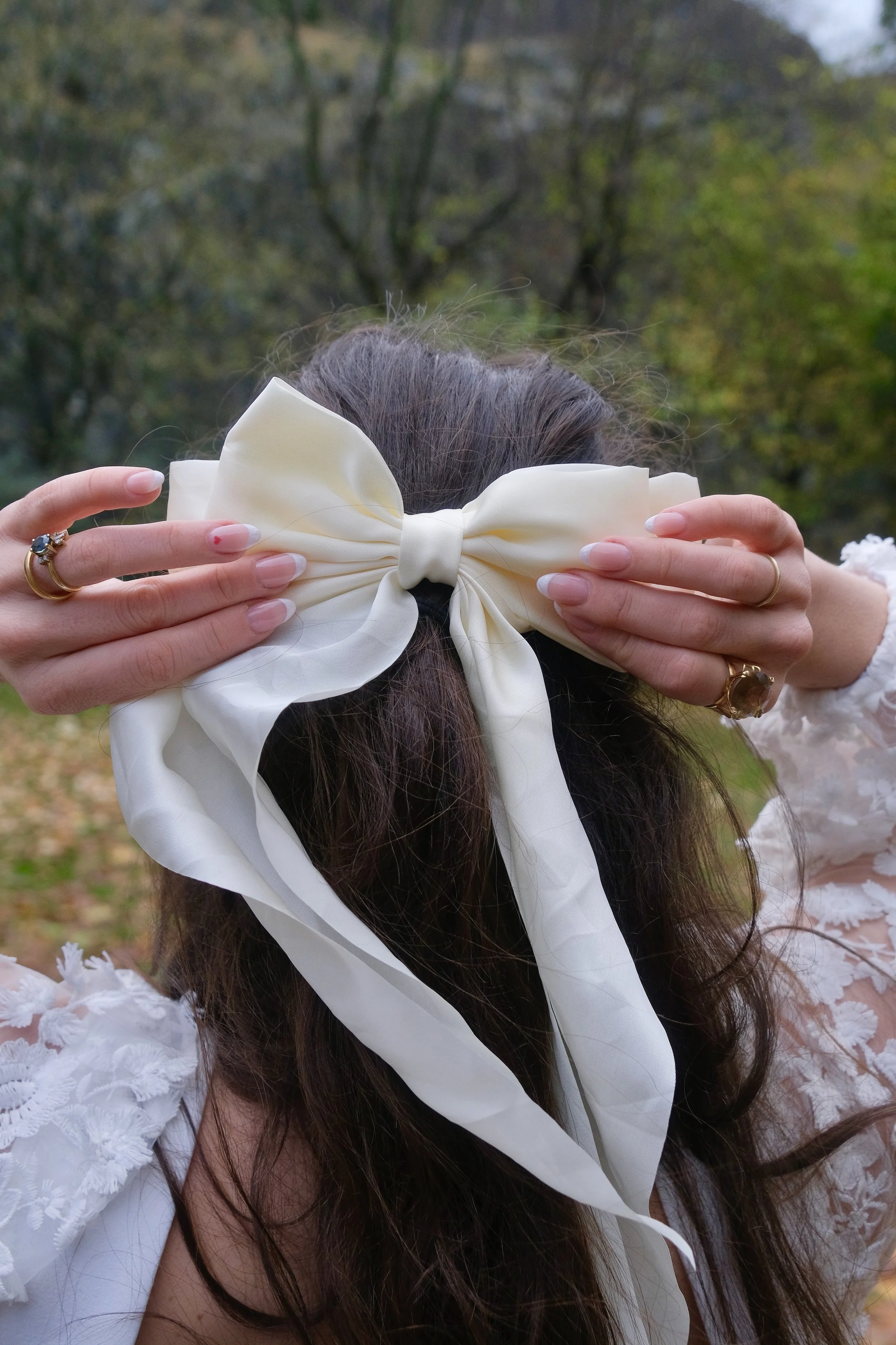 Person with long dark hair holding a large white bow over their face outdoors, wearing a white lace dress and multiple rings.