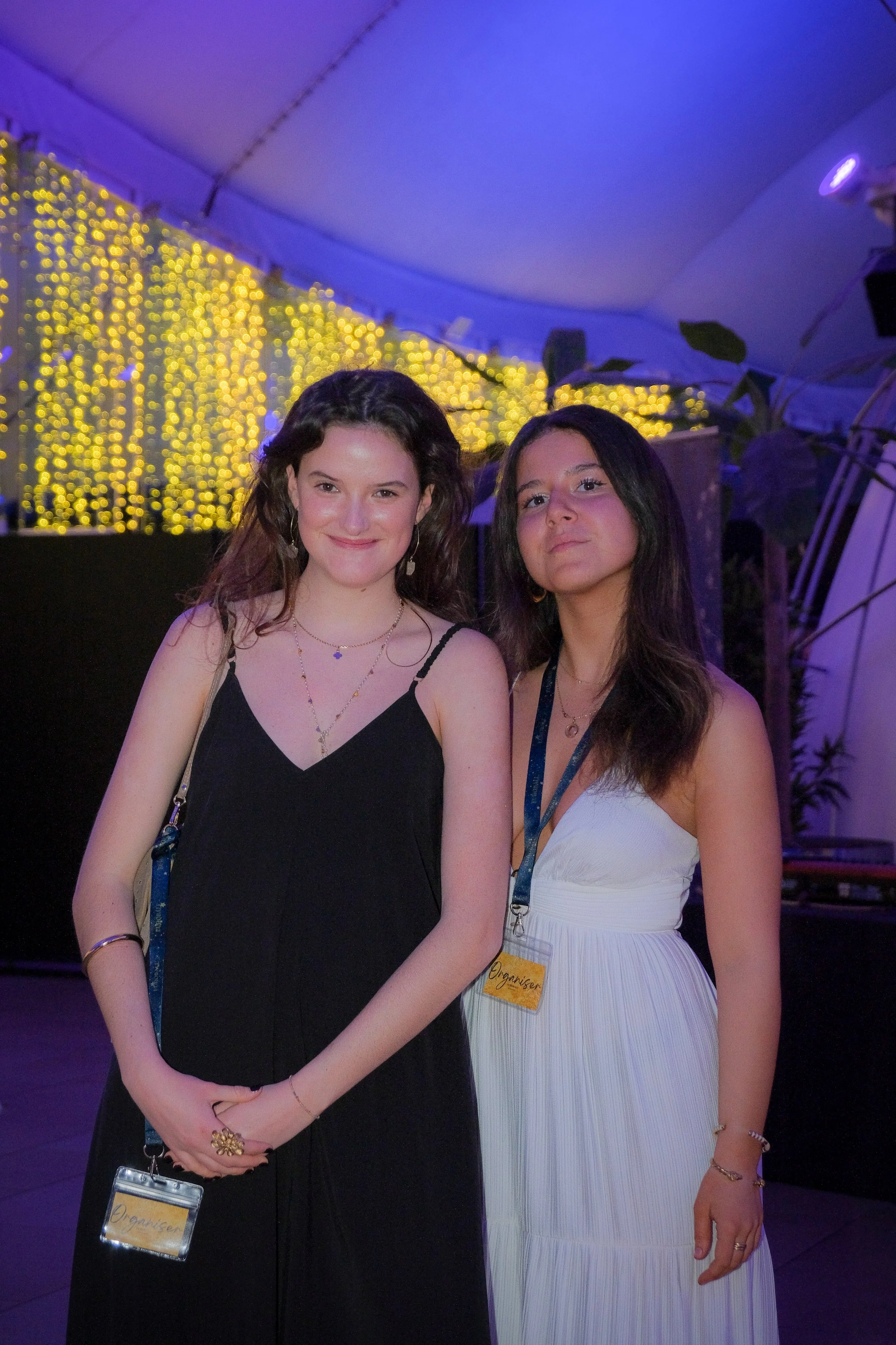 Two young women in dresses stand together at an indoor event with yellow string lights in the background.