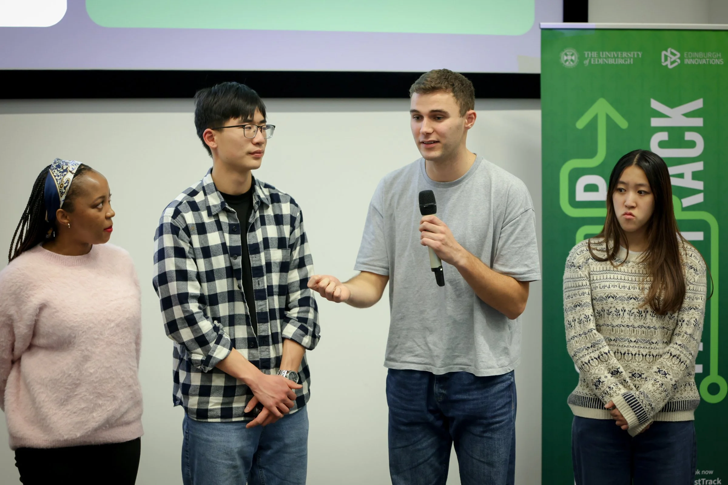 A group of four young people standing in front of a green banner with the words 'EDINBURGH' and 'STARTUP' and a logo, participating in a discussion or presentation, with one young man speaking into a microphone.
