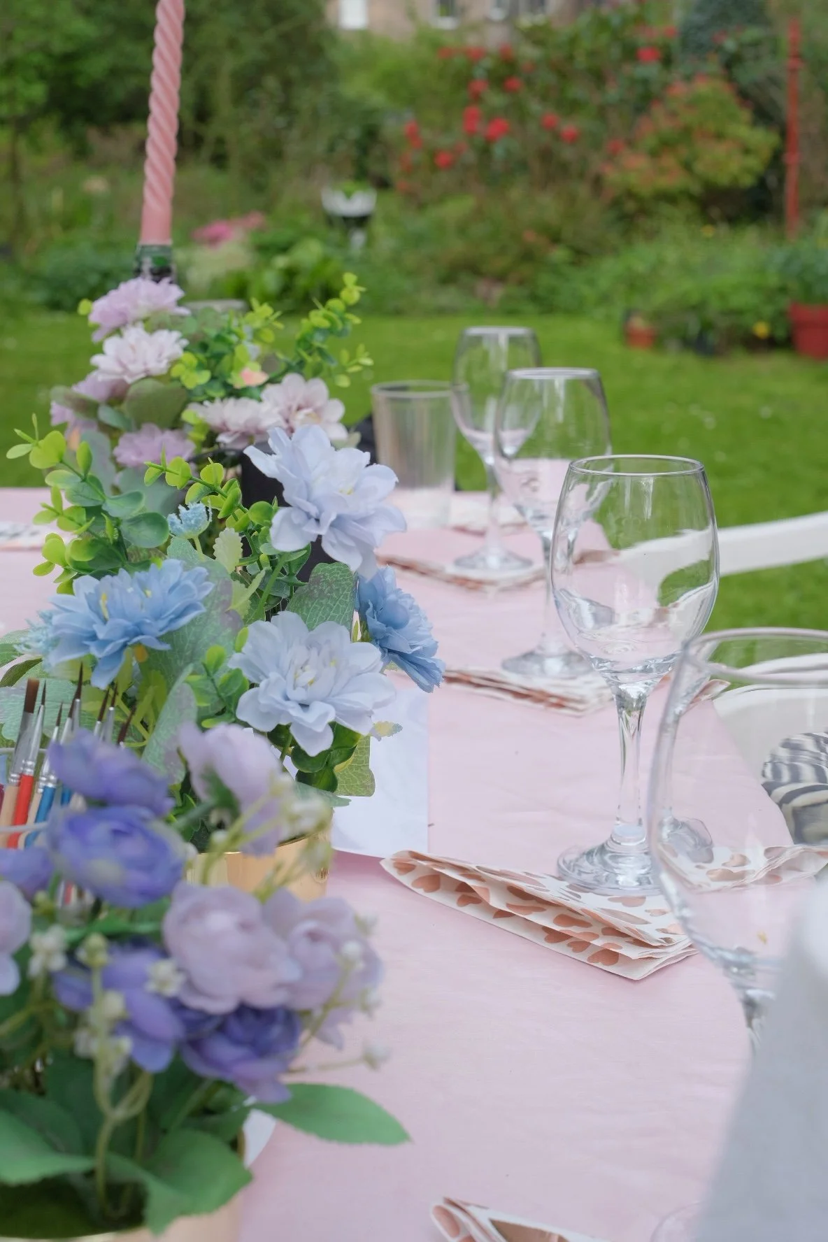 Table set outdoors with pink tablecloth, floral centerpiece, wine glasses, and napkins, in a garden with green grass and flowering bushes in the background.