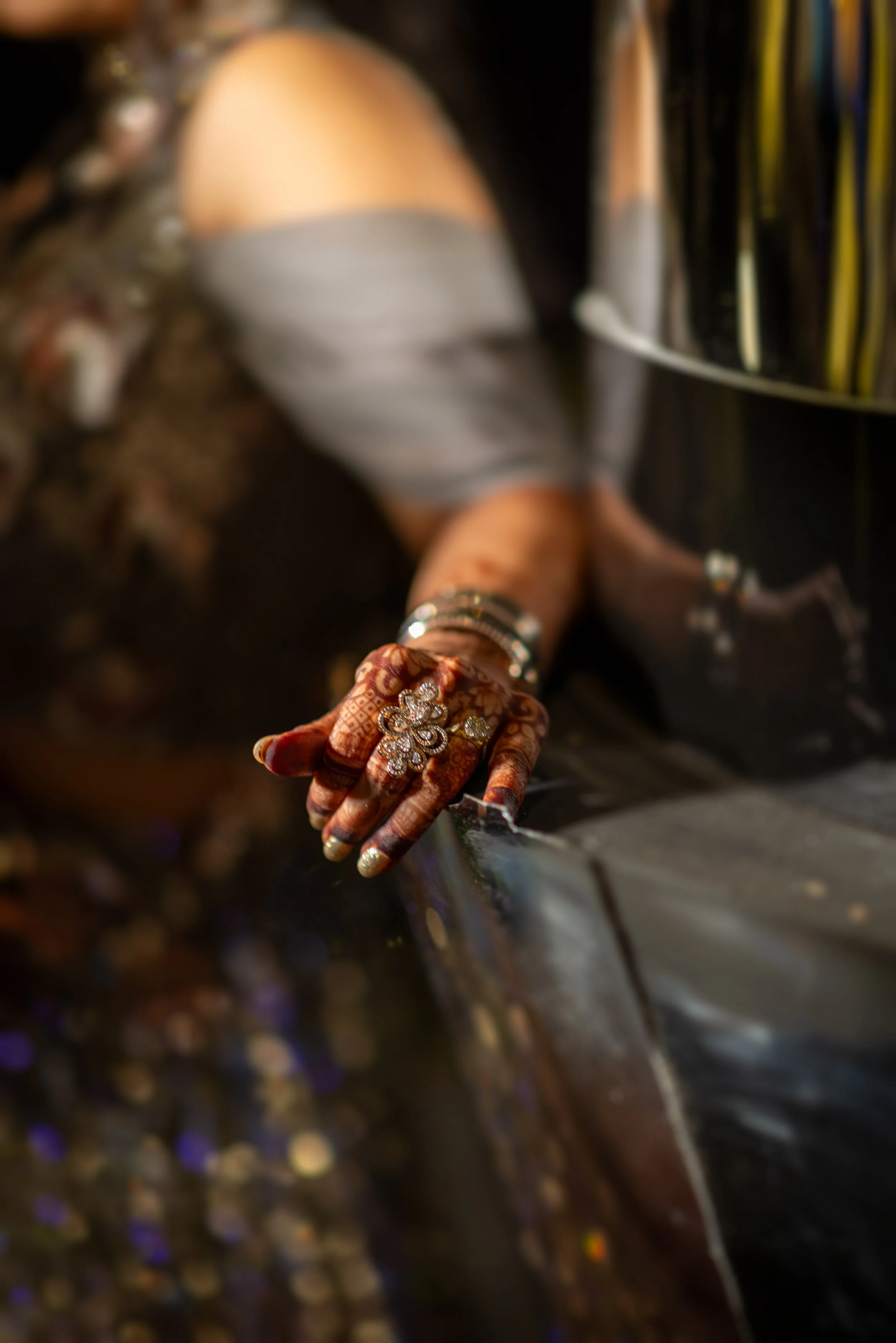 Close-up of a woman’s hand with decorative henna designs, wearing rings and bracelets, reaching out over a reflective, dark surface.