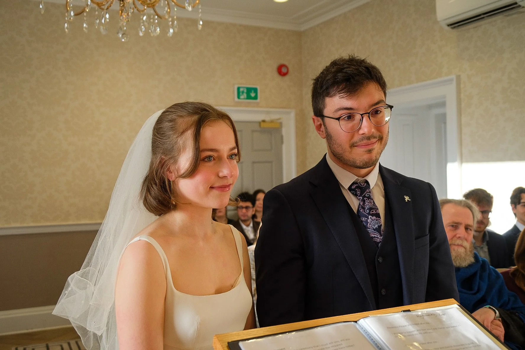 A bride and groom standing together during a wedding ceremony in a decorated indoor venue. The bride is wearing a white dress and veil, and the groom is dressed in a dark suit with glasses. Other seated guests are visible in the background.