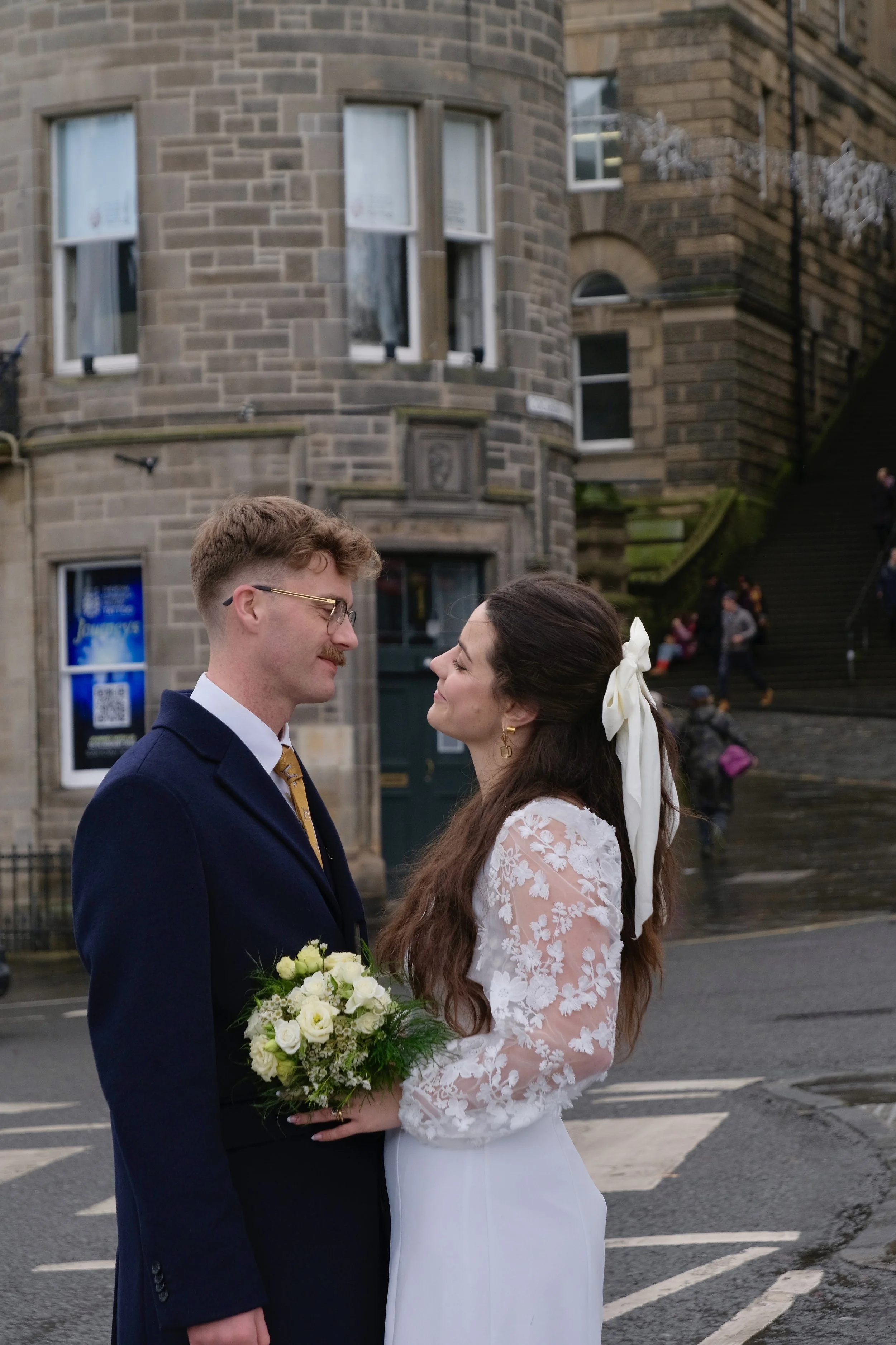 A bride and groom stand face-to-face holding hands on a city street, with the bride holding a bouquet of white flowers. The bride has long dark hair with a white ribbon in it, dressed in a white lace gown. The groom has short curly hair, glasses, and