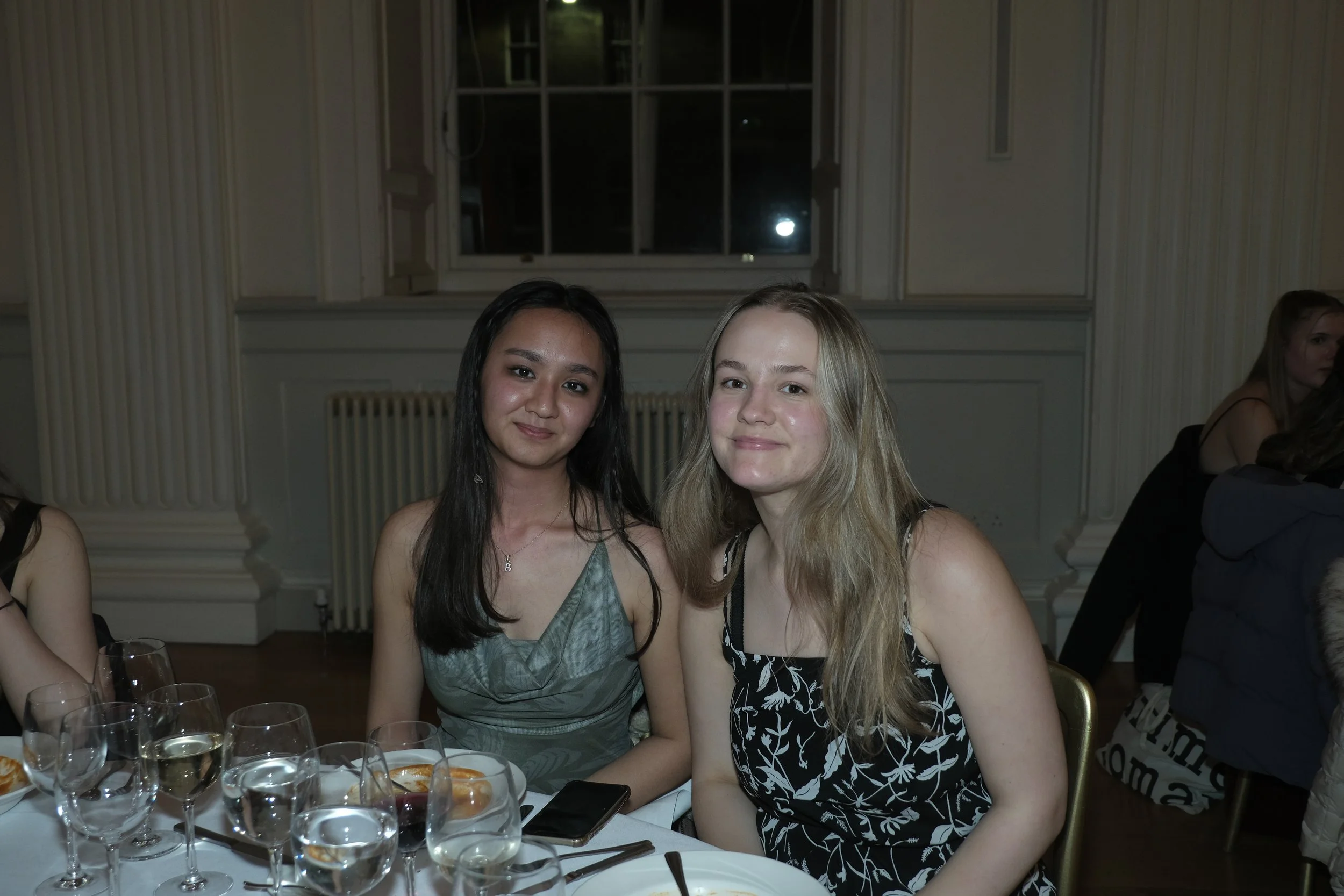 Two young women sitting at a formal dinner table with multiple wine glasses, dressed in elegant dresses, smiling at the camera in a well-lit and decorated room.