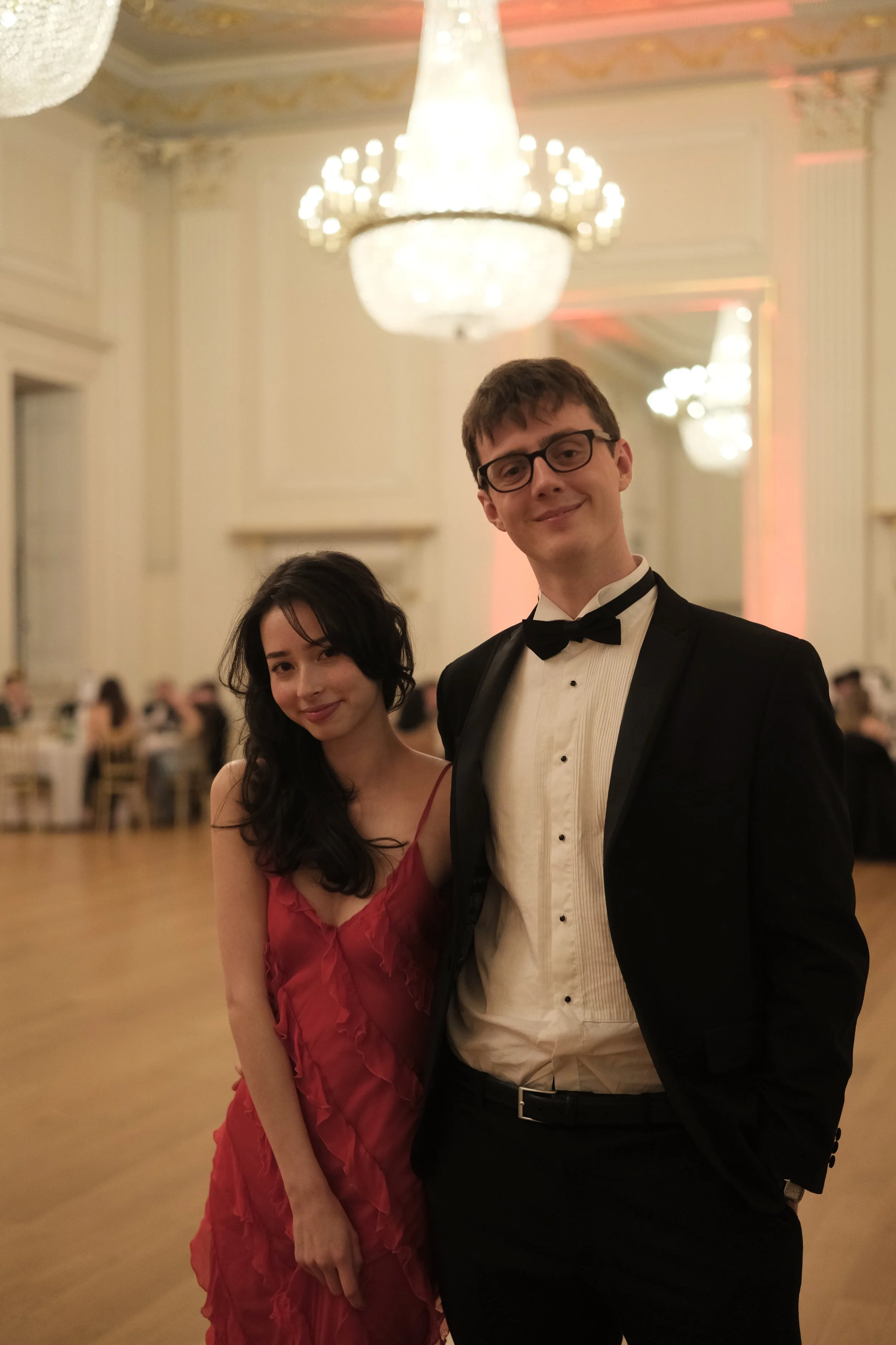 A young man and woman dressed formally at an elegant event in a ballroom with chandeliers and guests seated at tables.