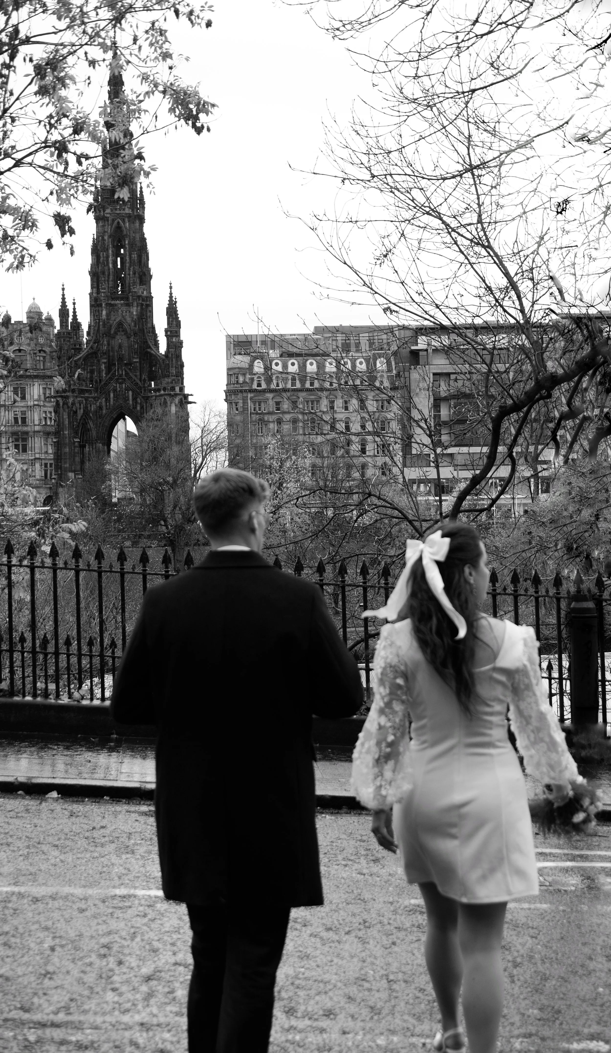 A black and white photo of a couple walking along a wet street, with historic buildings and a church tower in the background, and leafless trees surrounding a fenced park area.