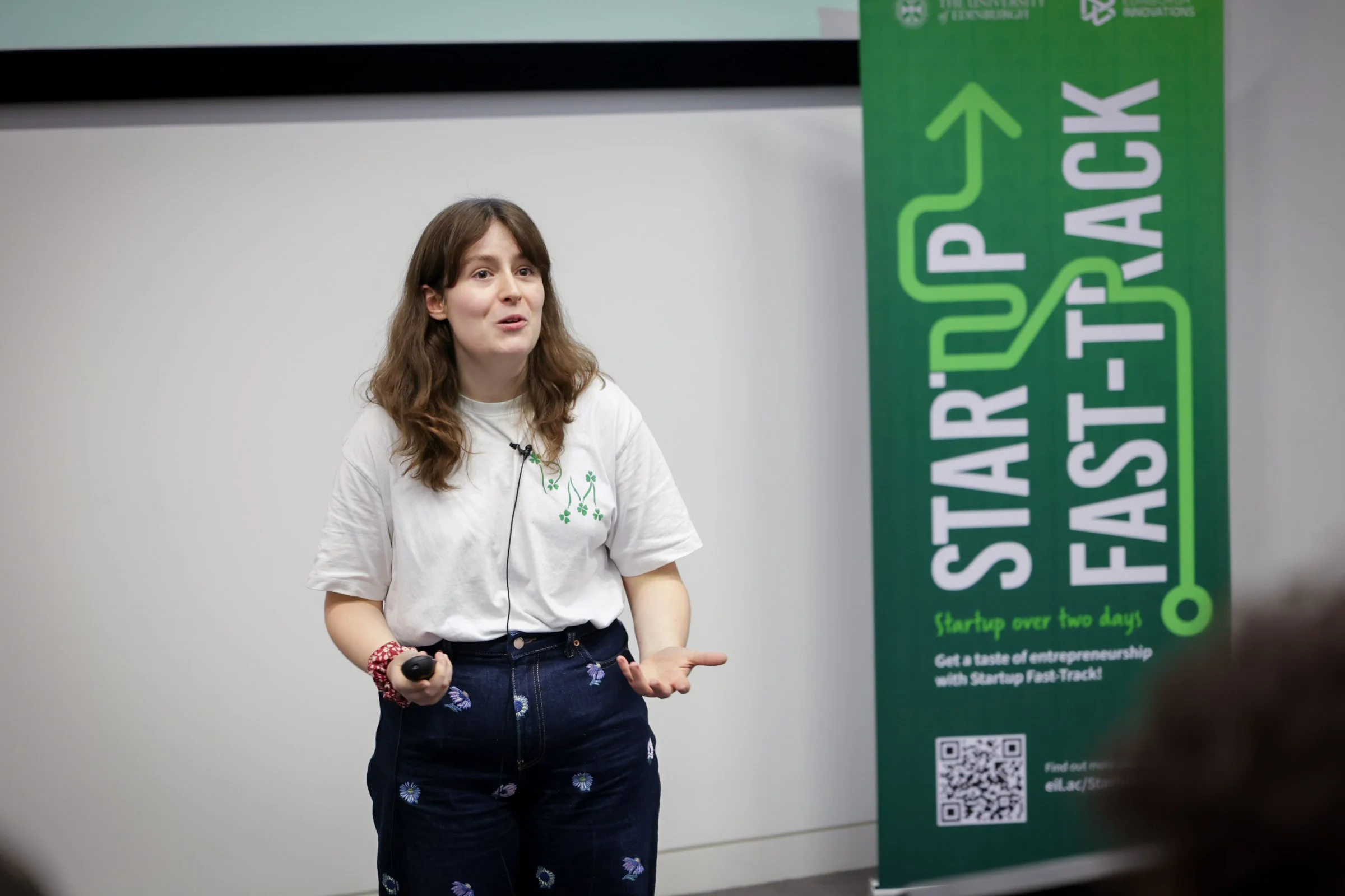 A young woman with brown hair, wearing a white T-shirt and dark blue pants with floral patterns, is giving a presentation indoors. She is holding a remote control and standing in front of a green banner with text related to Startup Fast-Track.