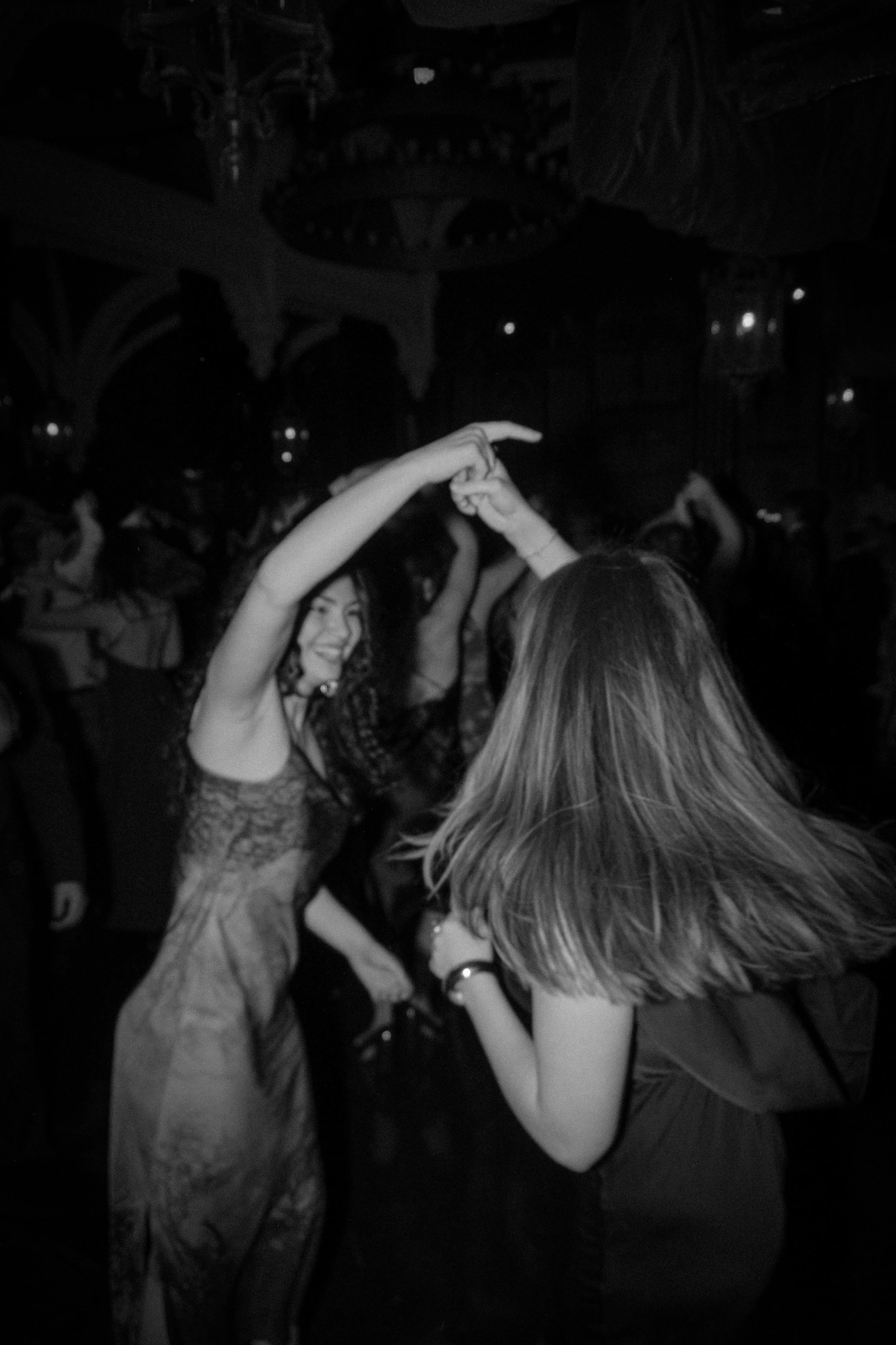 Two women dancing and holding hands at a party or nightclub with other people in the background.