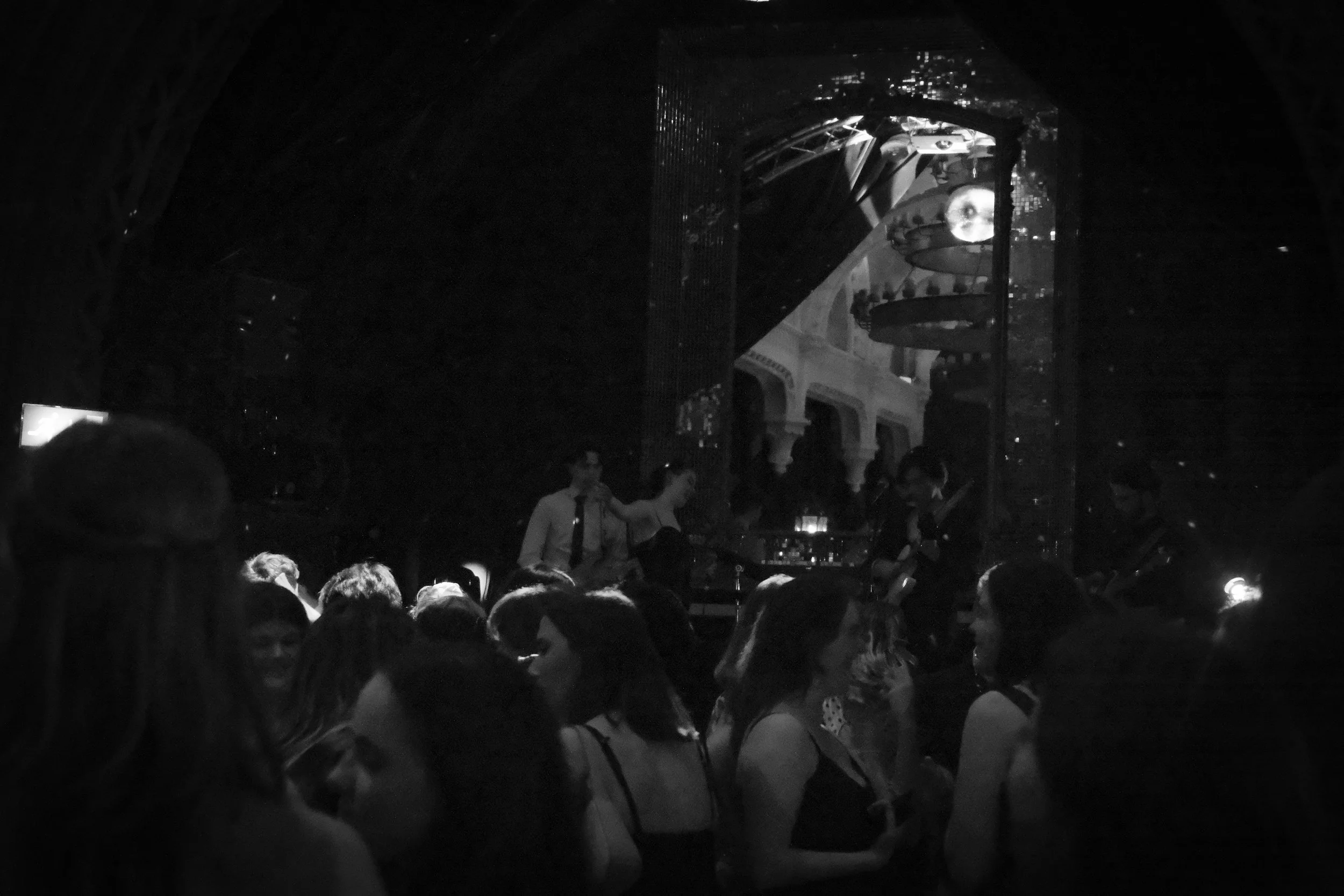 A crowded nightclub with people dancing and socializing. A stage with a band performing, visible through a large mirror on the back wall. Piano and other musical instruments on the stage. Dim lighting with some reflective surfaces.