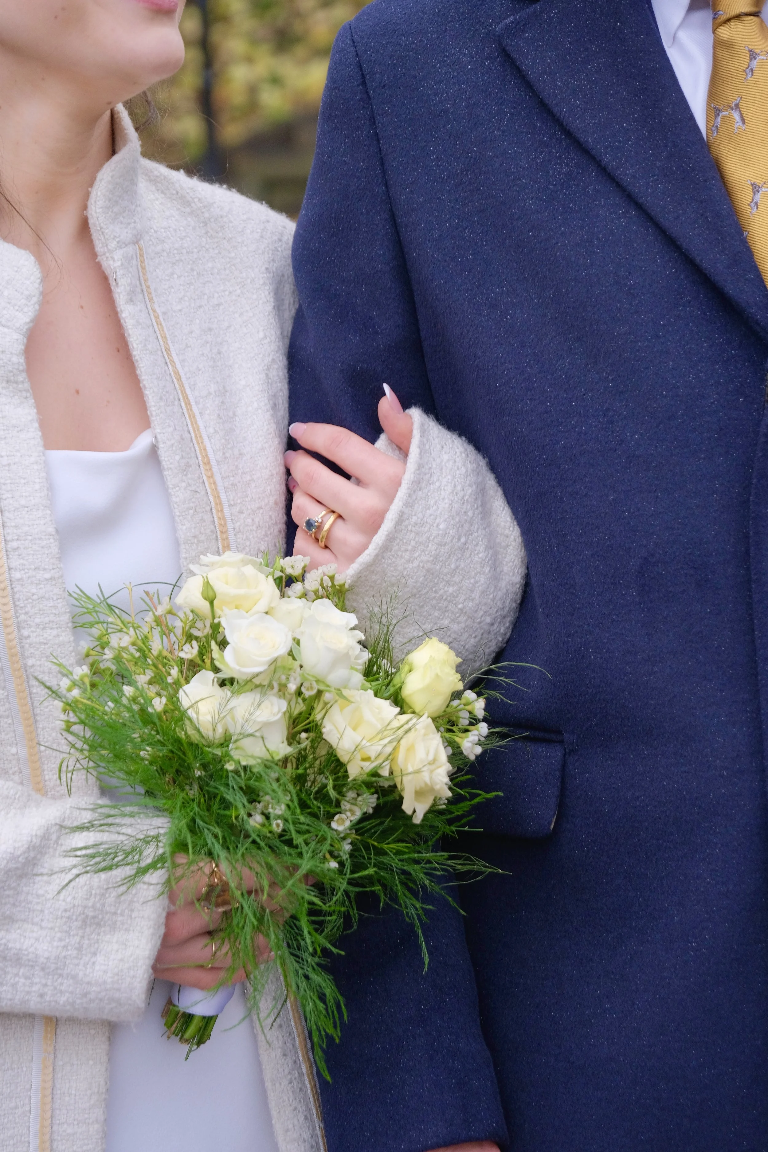 Close-up of a woman holding a bouquet of white roses and greenery, with an arm linked through a man's arm, both dressed in formal attire.