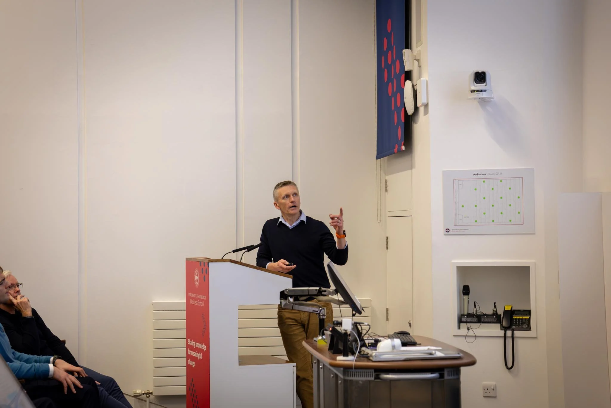 A man in a black sweater and khaki pants is giving a presentation at a university event, standing at a white podium with a university logo, in front of a white wall with a diagram of an auditorium layout, with some people sitting in the audience.