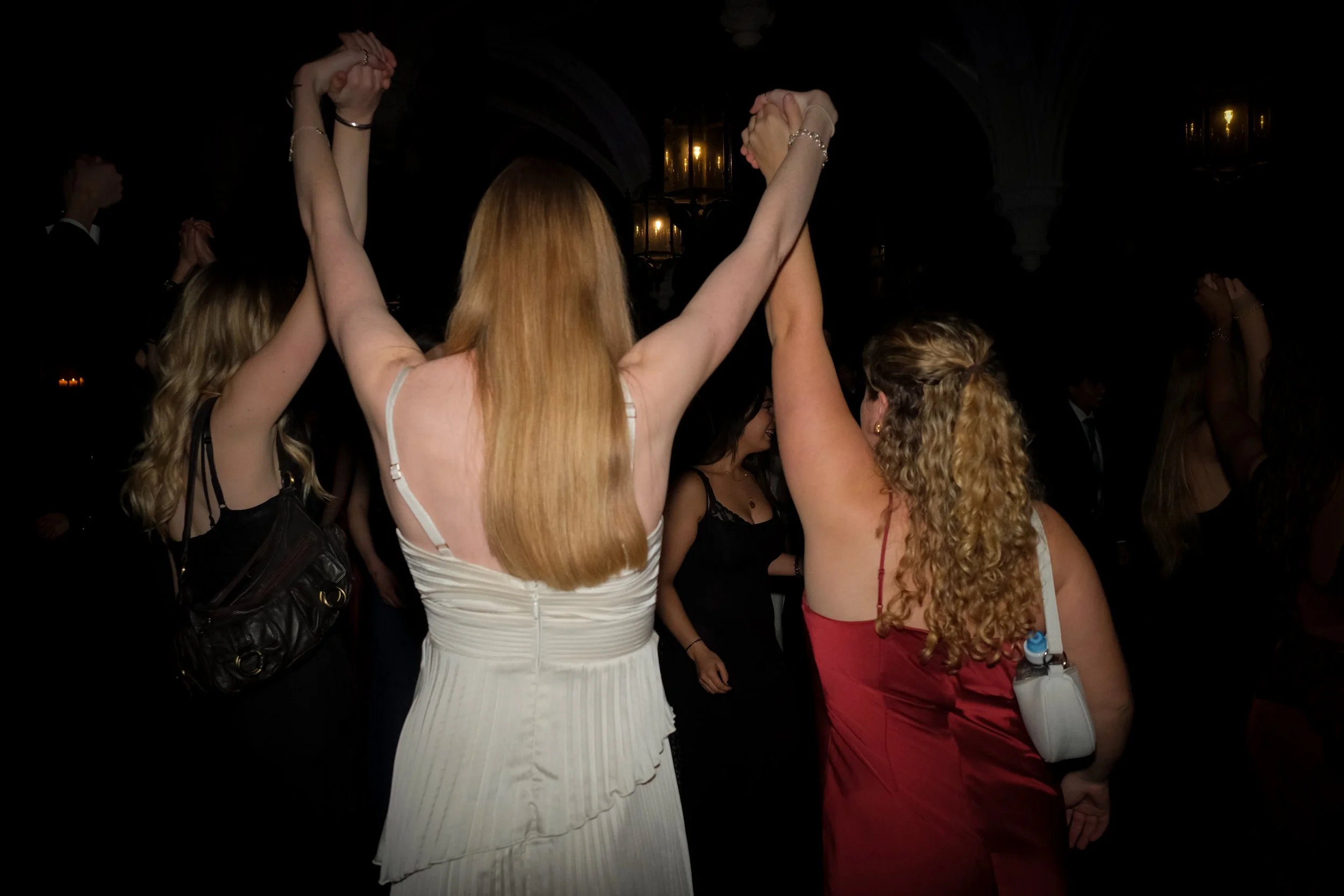 Two women with long hair dancing and holding hands up at a party or celebration in a dimly lit room.