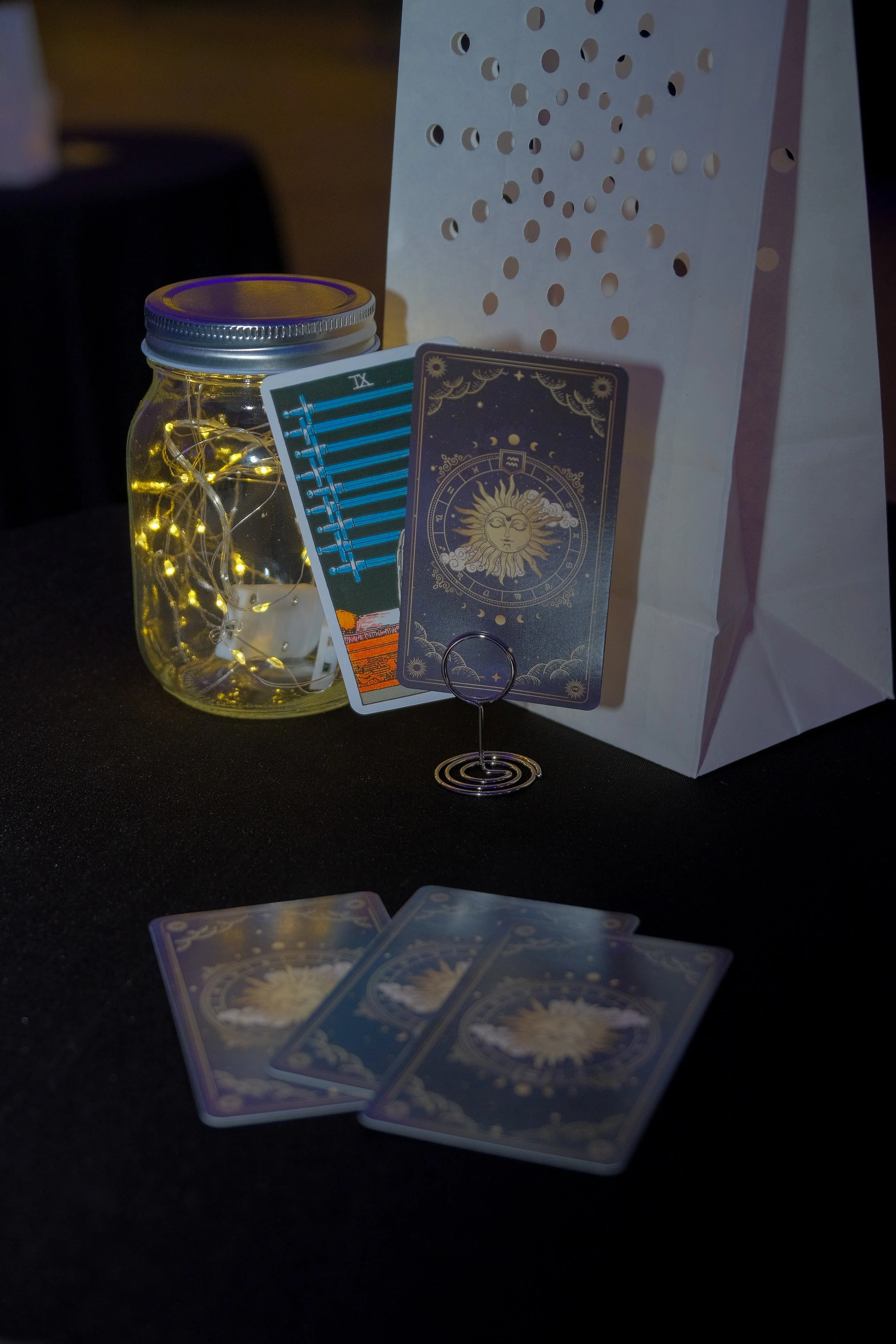 A jar with fairy lights, tarot cards, and a white bag with circular cutouts on a table.