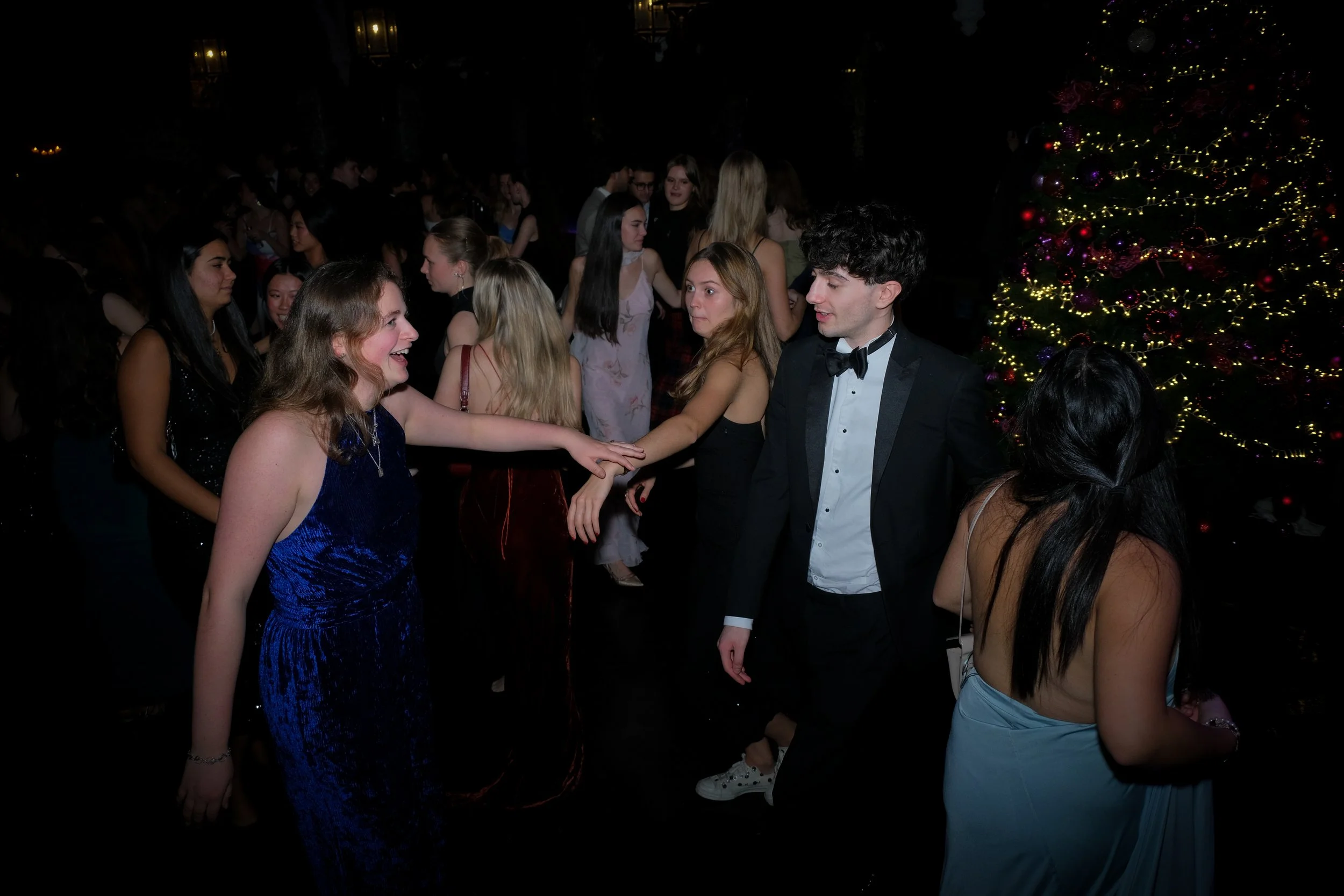 Young people dancing and socializing at a Christmas party with a decorated Christmas tree in the background.