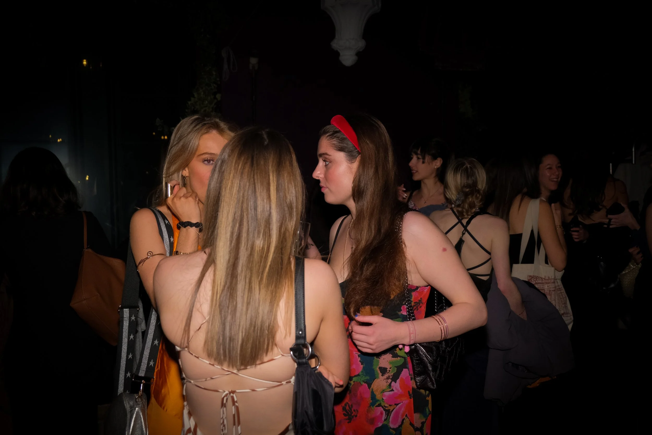 Group of women socializing at night, some engaged in conversation, women in casual and fashionable attire, dark background with dim lighting.