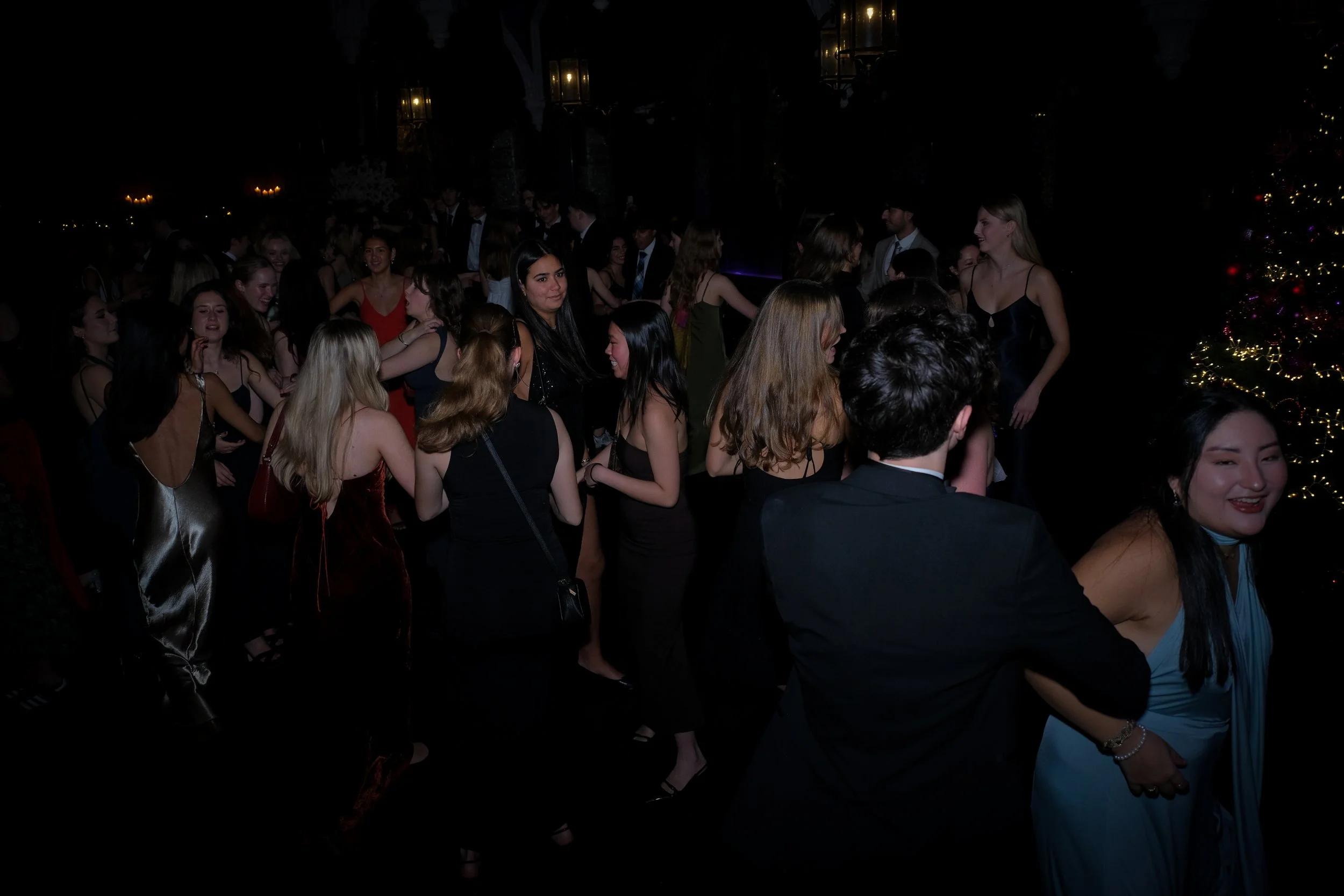 A group of people dressed in formal attire dancing and socializing at a party, with Christmas lights and a decorated Christmas tree visible in the background.