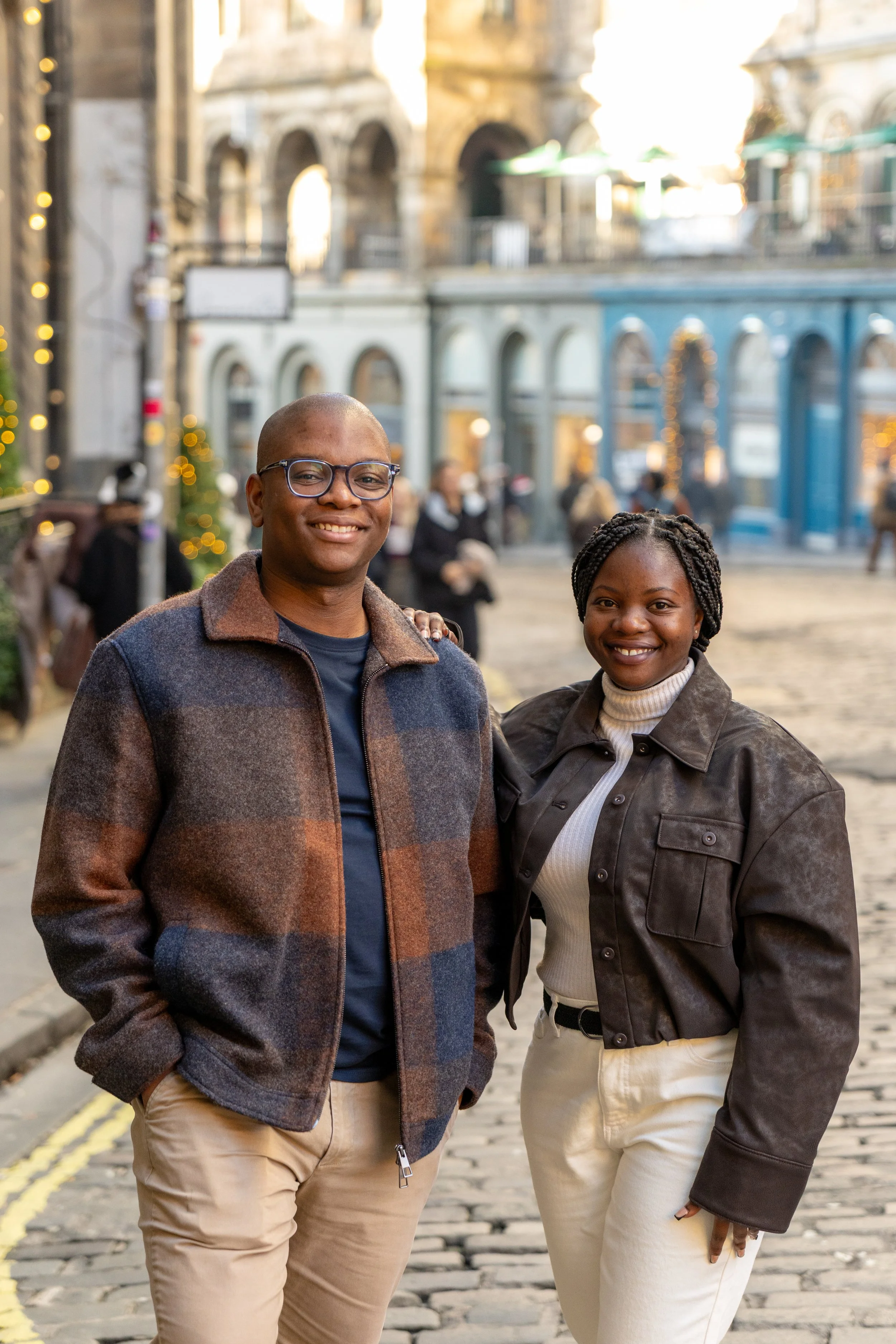 Two people standing together on a cobblestone street in front of historical buildings, smiling at the camera during the evening.