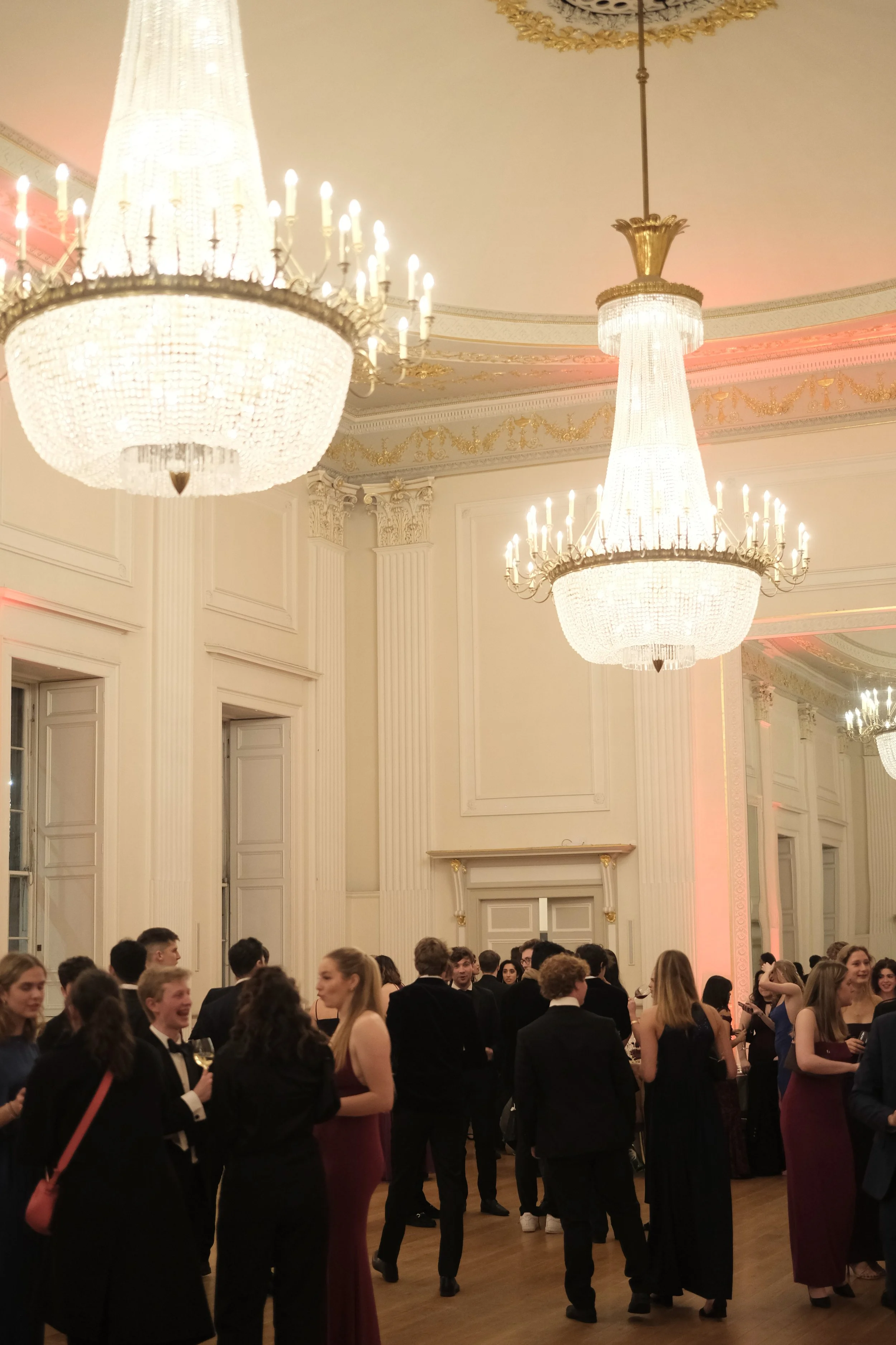 People dressed in formal attire socializing in an elegant ballroom with large chandeliers and ornate decor.