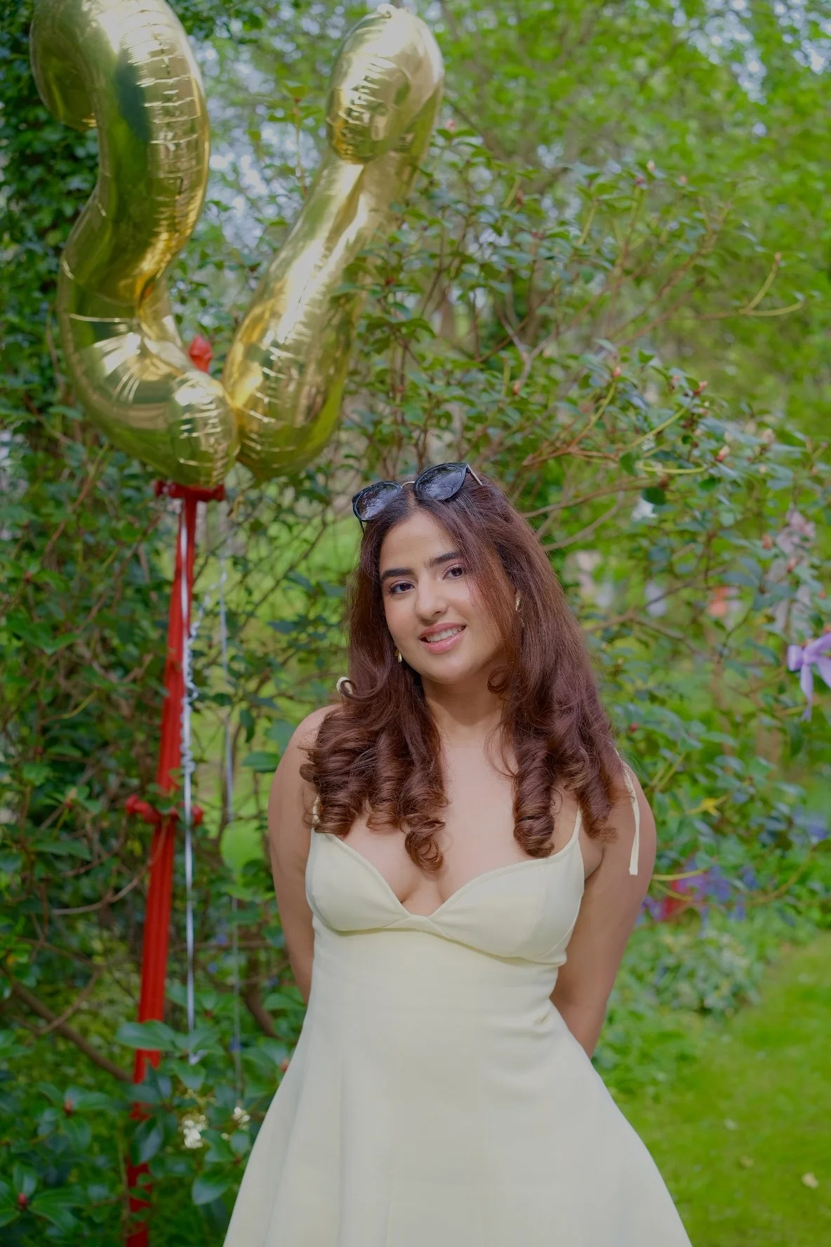 A young woman with long, curly brown hair, wearing a light yellow dress and sunglasses on her head, stands outdoors in a garden surrounded by green foliage. Behind her, gold foil balloons shape the number 23.