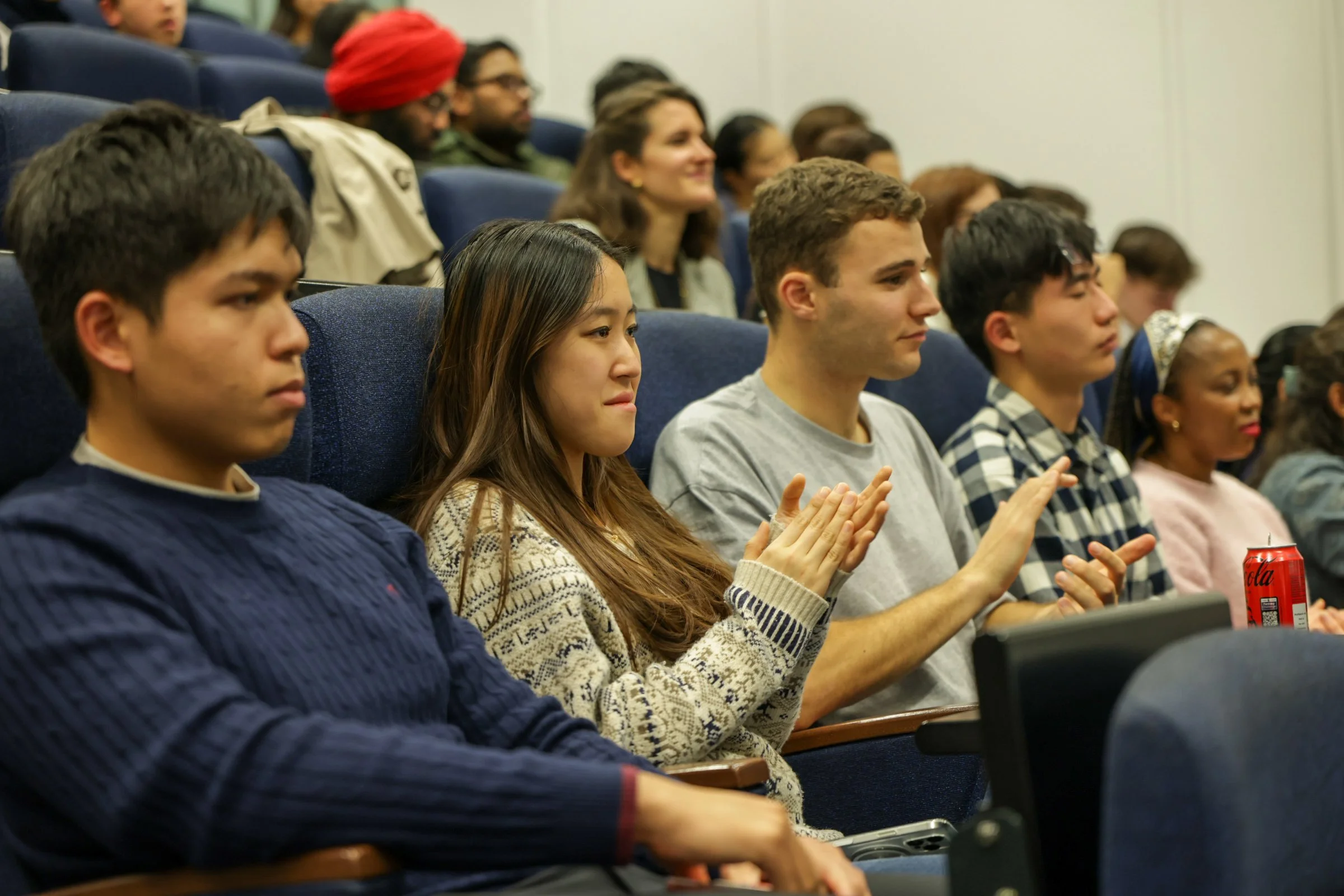 A diverse group of young adults seated in a theater or conference room, attentively watching an event or presentation, with some clapping and others focused.