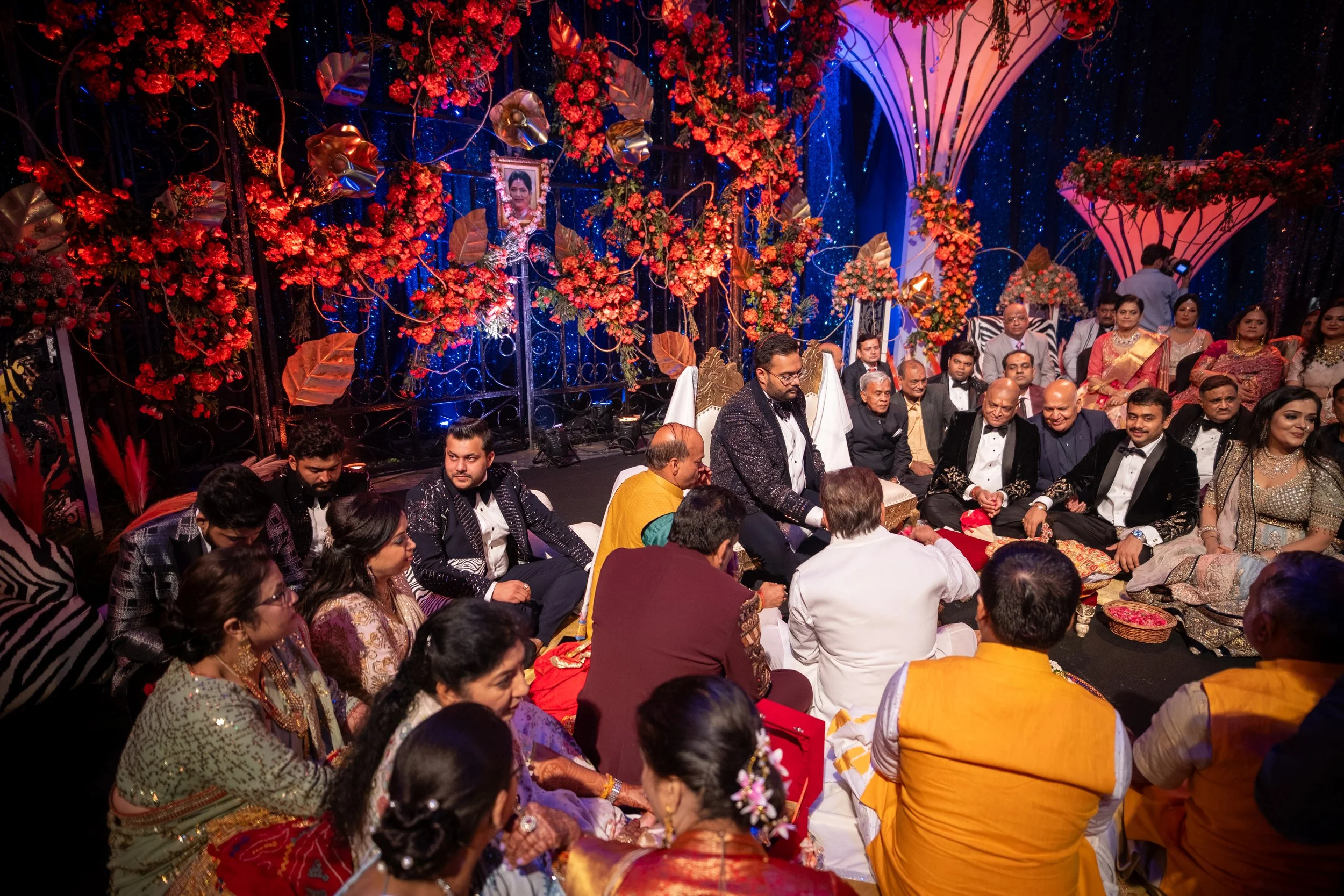 A large group of people attending an Indian wedding ceremony; the setting is decorated with vibrant red flowers, gold leaves, and blue drapery; the bride and groom are seated on ornate chairs at the center, surrounded by family and friends dressed in