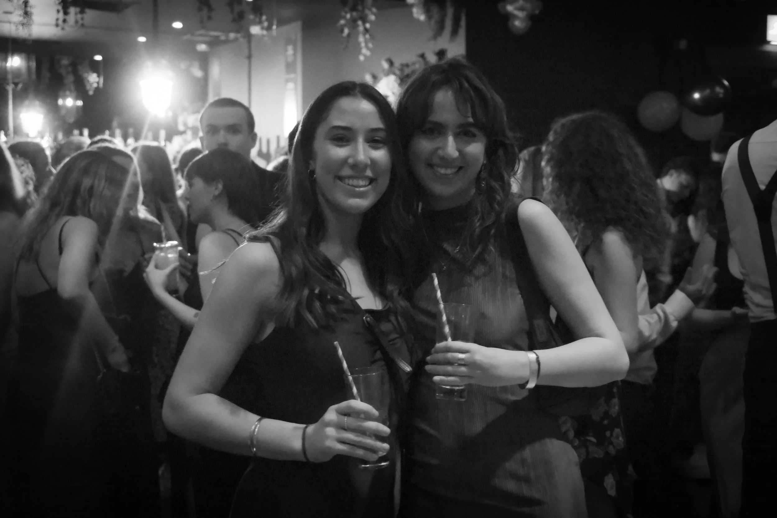 Two women smiling at a party, holding drinks, with a crowd in the background.