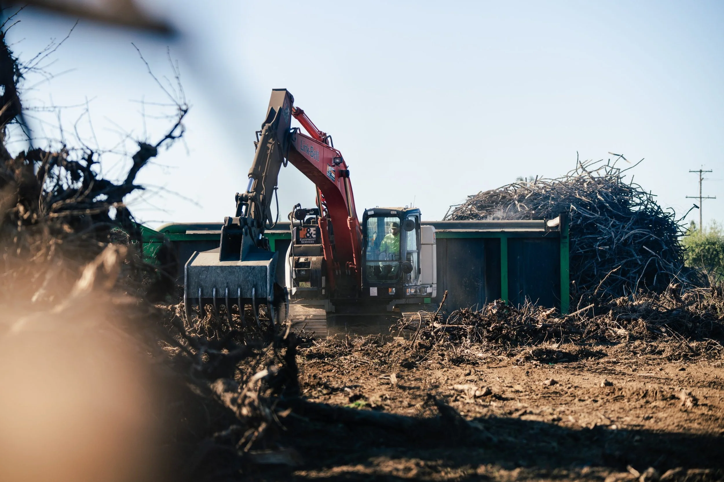 A bulldozer moving through a dirt field with piles of branches and debris, prepping for a airburner curtain air burning.