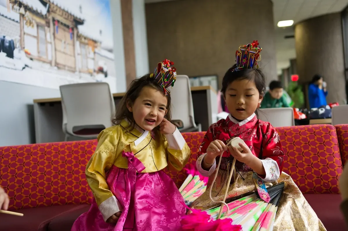 Two young girls in traditional Korean hanbok outfits, wearing colorful headpieces, sitting on a patterned red and orange couch inside a modern building, with other children in the background.