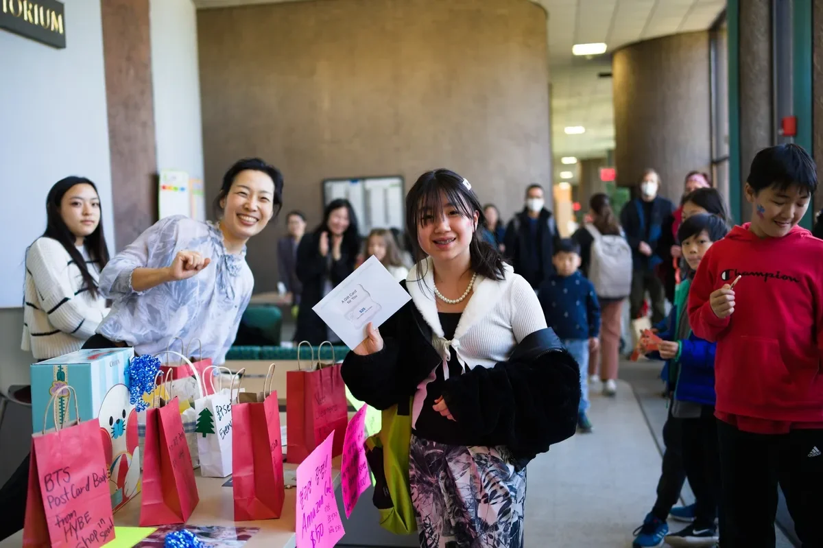 A young girl holding a letter or card at a table with gift bags, surrounded by smiling people in a busy indoor setting.