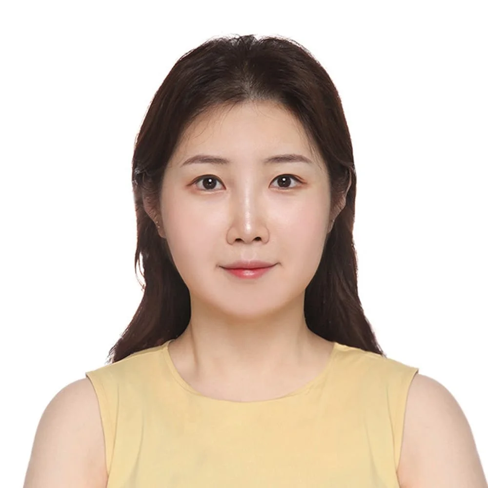 Portrait of a young woman with long brown hair wearing a yellow sleeveless top against a white background.