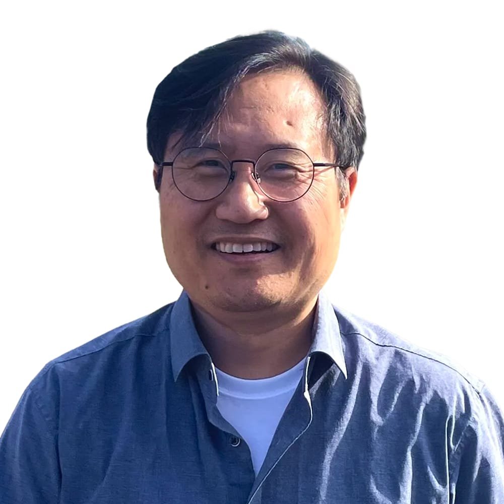 Smiling Asian man with glasses, wearing a blue shirt, standing outdoors in front of a plain white background.
