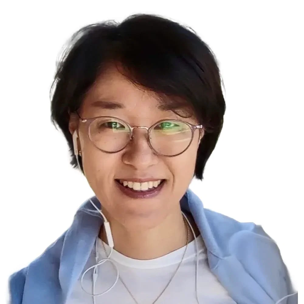 Smiling woman with short black hair, glasses, and earphones, wearing a blue hoodie over a white shirt, against a white background.
