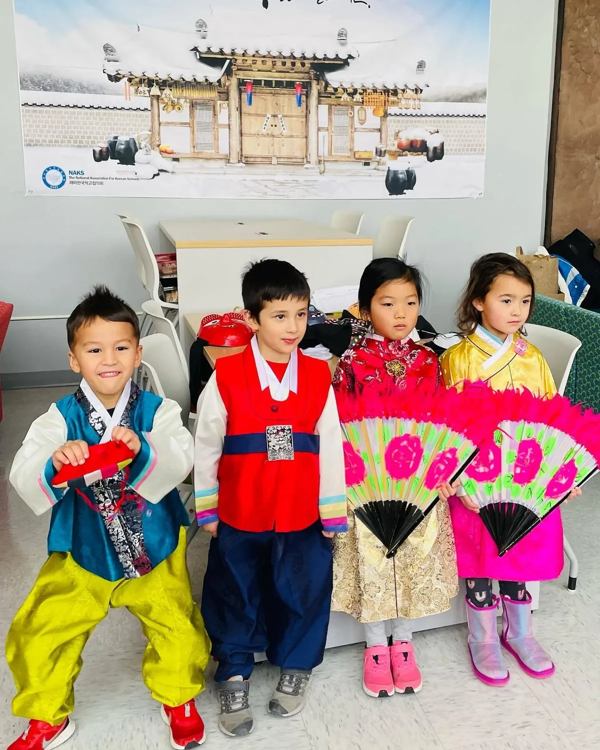Four children dressed in traditional Korean clothing, holding fans, standing indoors with a large poster of a traditional Korean house on the wall behind them.