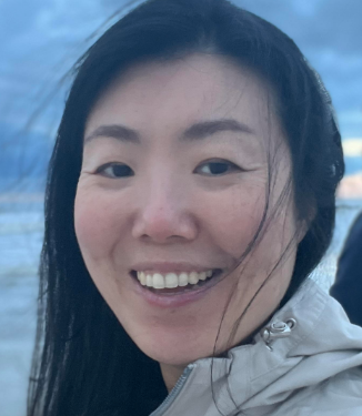 Close-up of a smiling woman with wet black hair, wearing a light-colored jacket, against a cloudy sky background.