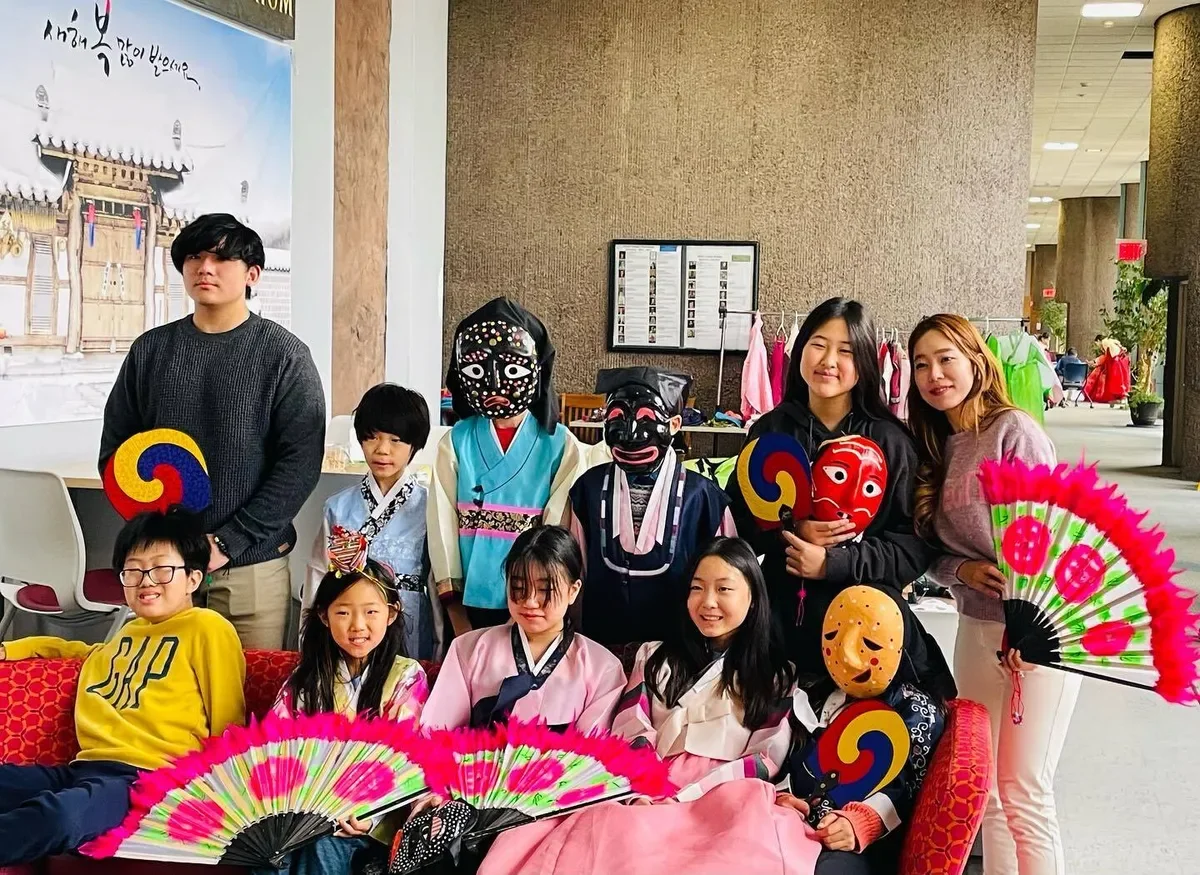 A group of people, including children and adults, in a cultural setting with traditional masks, fans, and colorful clothing, posing indoors in front of a historical painting and a display of costumes.