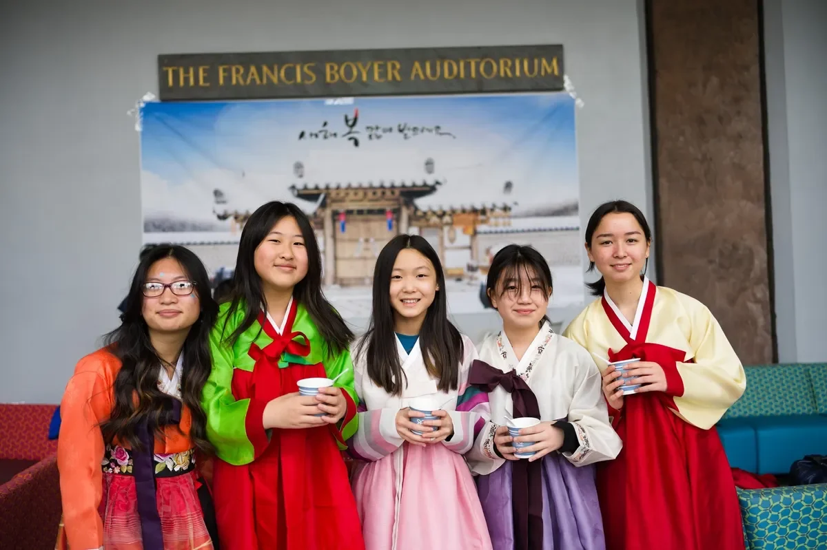 Five young women standing in front of a sign that reads "The Francis Boyer Auditorium"; they are wearing traditional Korean hanbok dresses and holding cups, smiling for the camera.