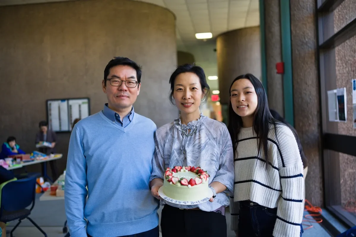 Three people posing with a cake in an indoor space, likely a celebration, with other people and tables in the background.