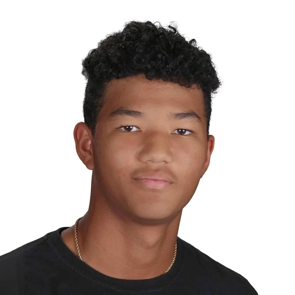 Portrait of a young man with curly black hair wearing a black shirt and a gold chain, against a plain white background.