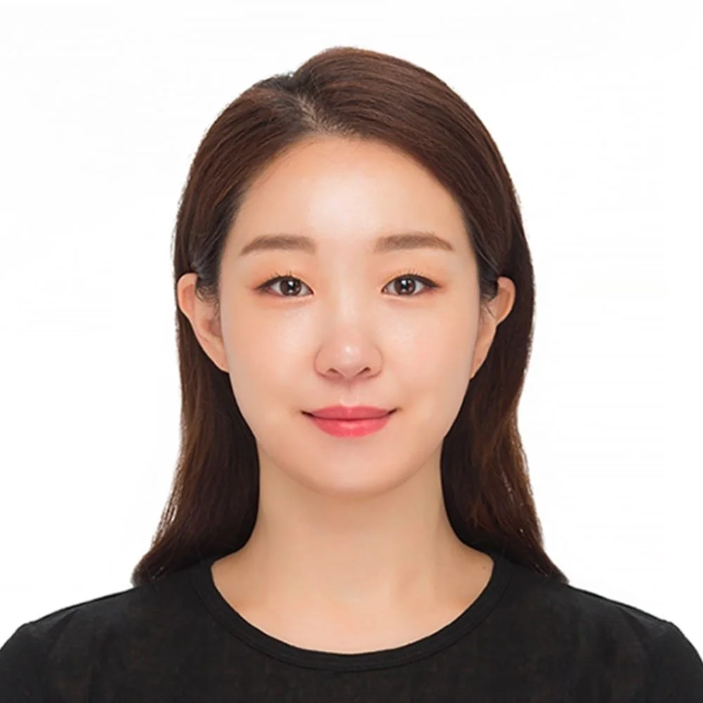 A woman with shoulder-length dark brown hair, light skin, wearing a black top, smiling, against a white background.