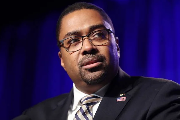 Thomas Gary with glasses wearing a suit and tie, with an American flag pin on his lapel, against a dark blue background.